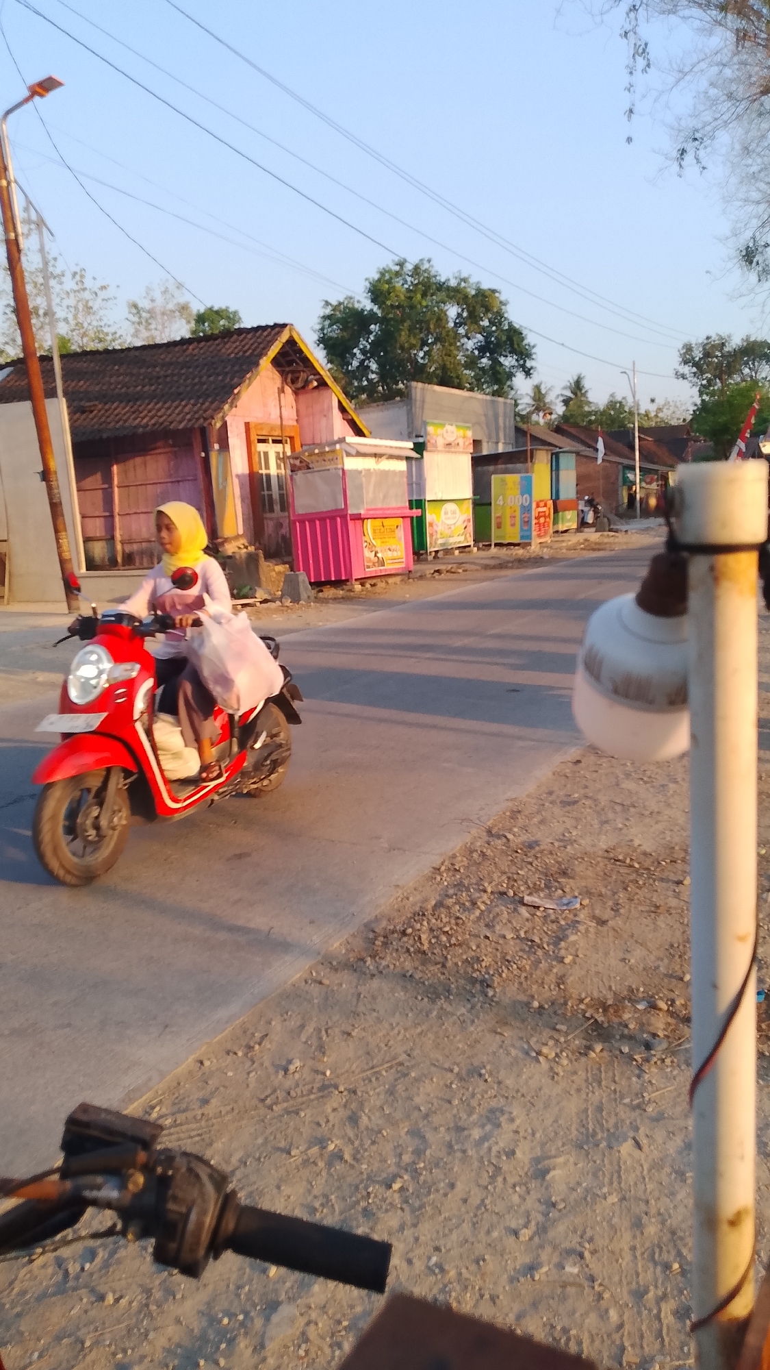 A woman wearing a yellow hijab and a light pink shirt rides a red scooter along a paved road in Karanglangu, Indonesia. She is carrying a large, translucent plastic bag filled with items on the back of the scooter. The scooter's headlight is on. She is traveling past a row of small buildings that appear to be shops or food stalls. The buildings have brightly colored fronts, with one pink stall prominently displaying a sign with images. A lamp post with wires is visible on the left side of the image, and another white pole with a white light fixture attached is in the foreground on the right. The road surface is asphalt, with a gravel and dirt shoulder on the right. The sky is clear and blue, indicating daytime. The sun casts long shadows, suggesting it is either early morning or late afternoon. The overall scene depicts a typical street view in a semi-urban or rural area of Indonesia.