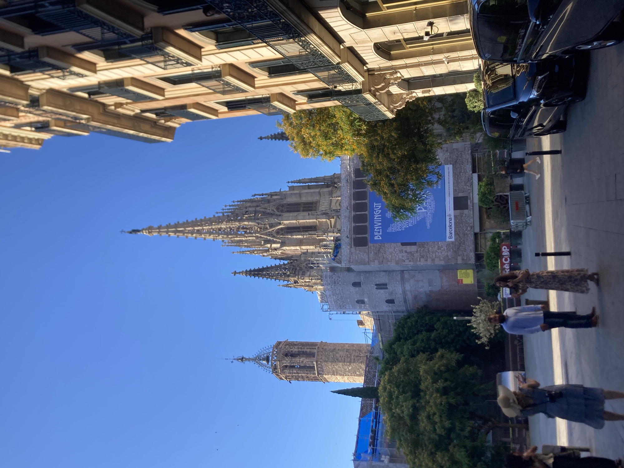 The image depicts a street scene in Barcelona, Spain, dominated by the architectural grandeur of the Barcelona Cathedral. The cathedral's intricate Gothic spires and a tall bell tower are prominently visible against a clear blue sky. The foreground features an ornate building with balconies, typical of Catalan architecture. In the middle ground, there are several people. A woman in a blue dress and wide-brimmed hat is taking a photo with a camera. Beside her, a man in a light blue shirt and jeans stands with a woman in a long, floral dress who is speaking. Further in the background, a large blue banner with text in Catalan, including "BENVINGUT" and "Barcelona," is displayed on the cathedral. Two black vans are parked on the right side of the street. Lush green trees, some with autumn foliage, are interspersed throughout the scene, adding a touch of nature to the urban landscape. The overall impression is one of a sunny day in a historic city.
