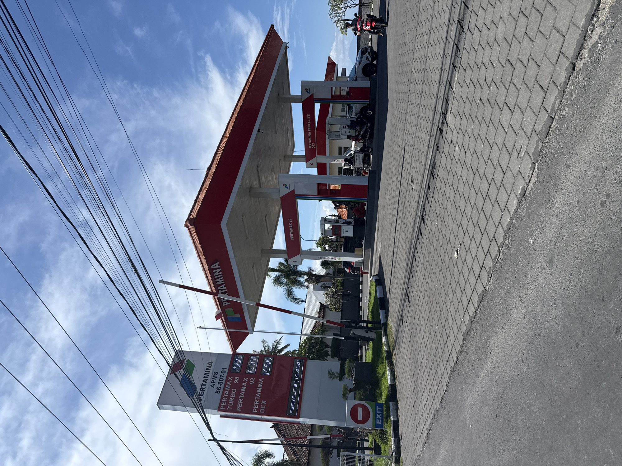 The image shows a Pertamina gas station in Batununggal, Indonesia. The gas station has a red and white canopy with Pertamina branding. A sign displays fuel prices for Pertamax Turbo 98, Pertamax 92, and Pertamina Dex. An "EXIT" sign is visible near the station entrance. A brick-paved street runs adjacent to the station, with parked motorcycles. Overhead, power lines are present against a blue, cloudy sky.