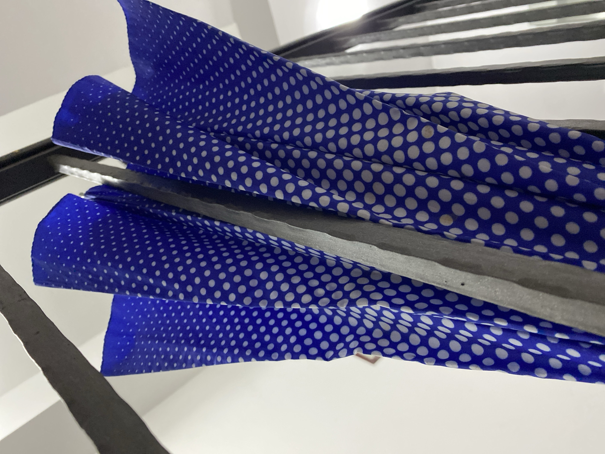 The image depicts a close-up, low-angle view of a blue polka dot scarf draped over a metal structure, likely a staircase railing. The fabric has a vibrant blue background with white polka dots evenly spaced across its surface. The metal structure appears to be dark-colored, possibly black or dark gray, with a metallic sheen. The background consists of a white or light-colored surface. There is no indication that this photo was taken in Budva, Montenegro.