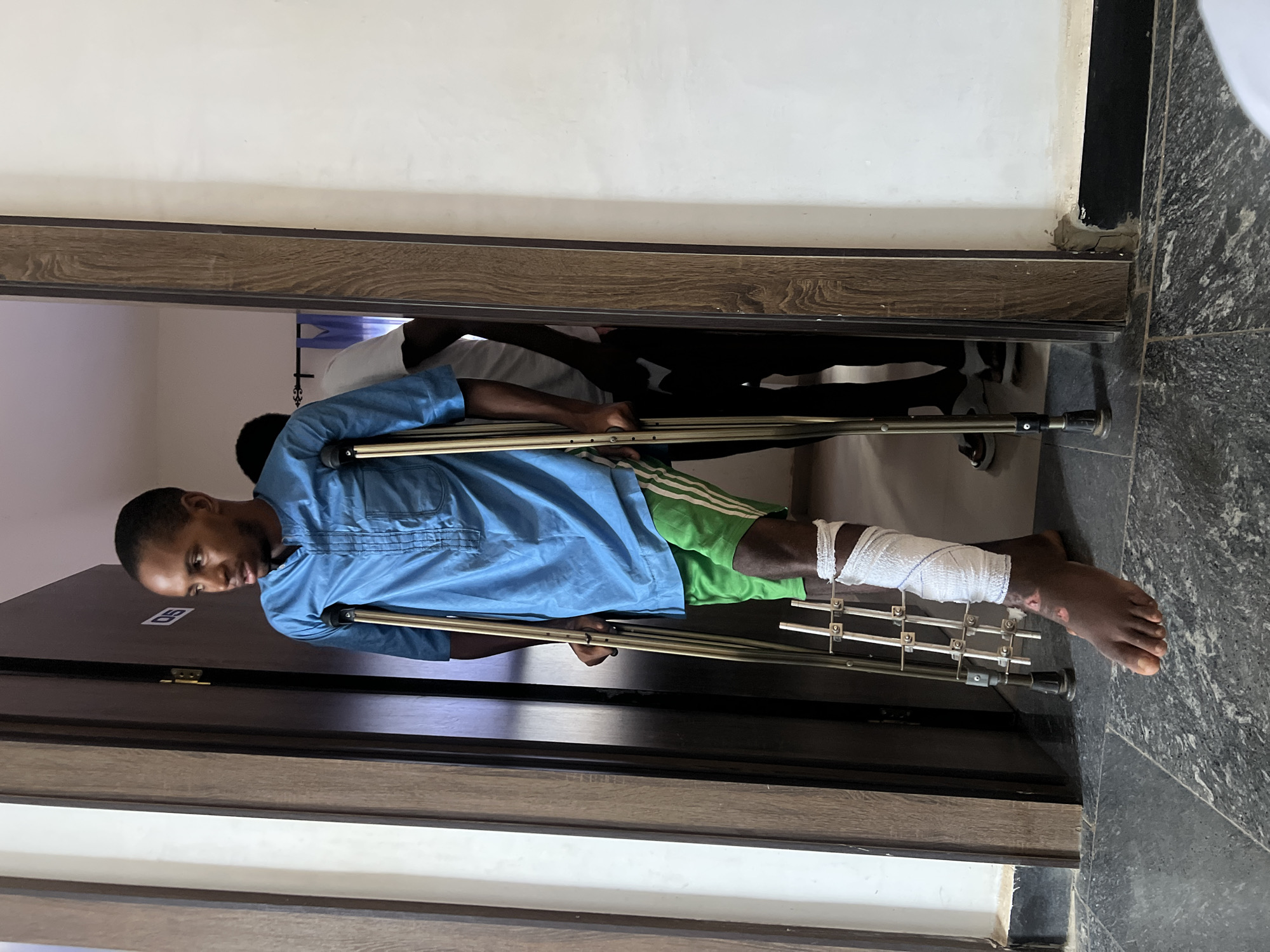 A man with a bandaged leg is using crutches to navigate through a doorway. The man is Black, wearing a blue button-down shirt and green Adidas shorts. His left leg is bandaged and has an external fixator attached. He appears to be indoors. The background shows a glimpse of other people in what looks like a hospital setting. The location is Jos, Nigeria. The floor is a dark, speckled stone.