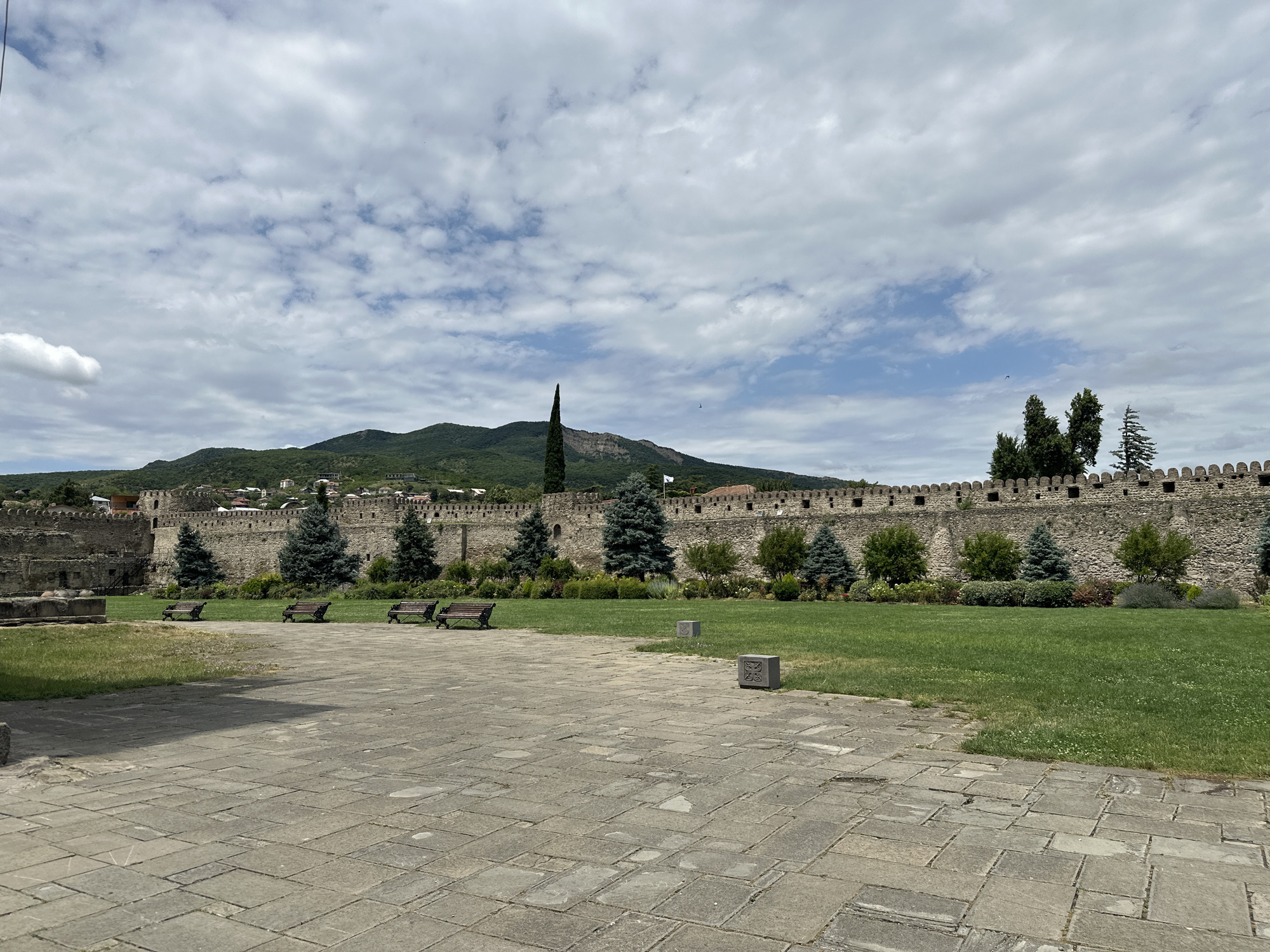The image shows a view of the Svetitskhoveli Cathedral complex in Mtskheta, Georgia. The foreground is a stone-paved courtyard. To the left, a section of ancient stone wall, part of the cathedral complex, is visible. A green lawn stretches across the middle ground, lined by a long, imposing stone wall with battlements. Several wooden benches are placed on the lawn. Behind the wall, a town with red-tiled roofs can be seen nestled at the foot of a green, forested mountain range under a cloudy sky. Tall, slender cypress trees and smaller evergreen trees are planted along the lawn in front of the wall.