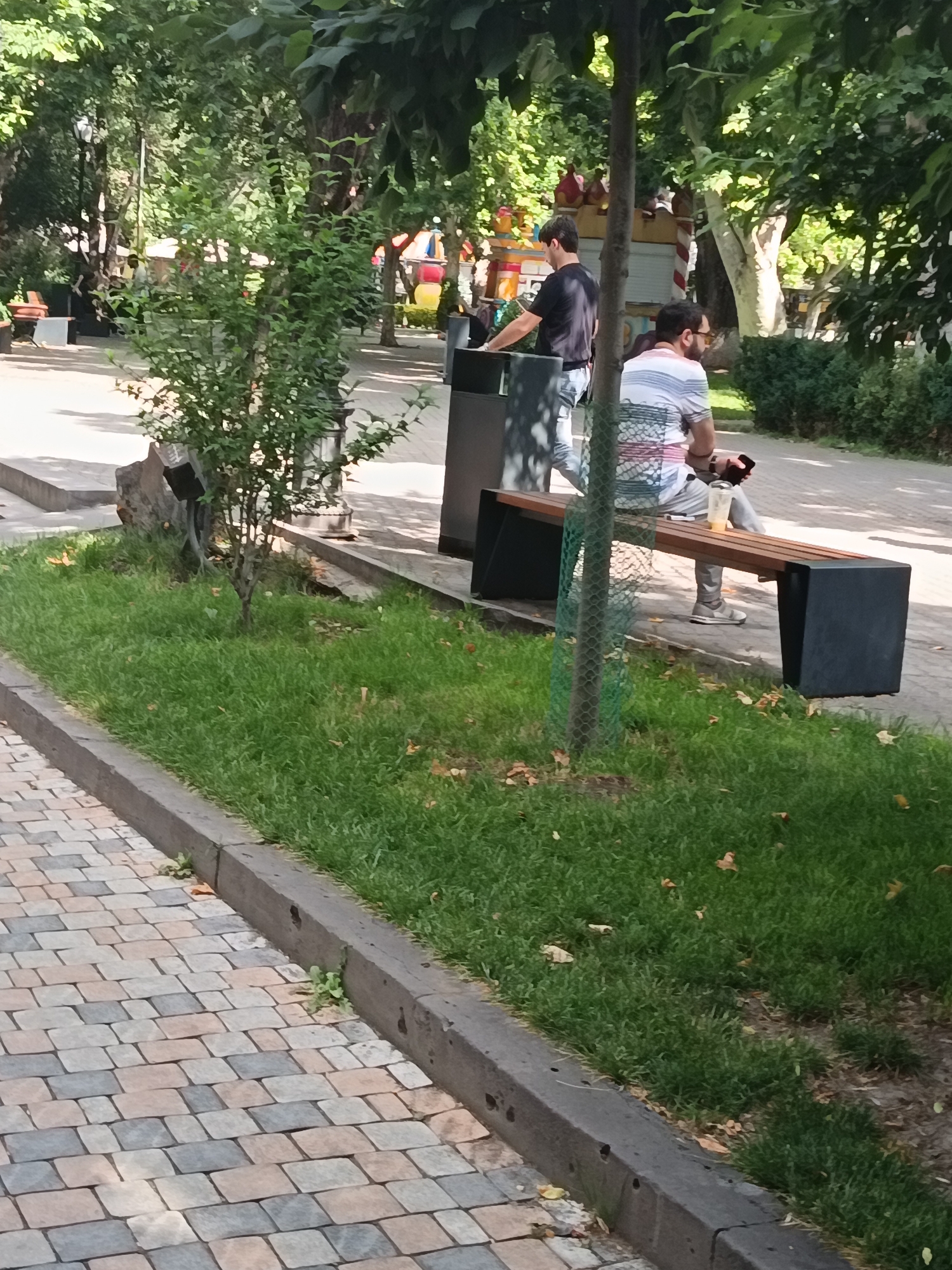 Two men are in a park setting. One man, wearing a black t-shirt and light-colored pants, is standing near a trash receptacle. He is leaning forward, possibly interacting with the receptacle. The second man, dressed in a striped t-shirt and grey pants, is seated on a park bench. He is looking to his right, holding a mobile phone in his left hand and a plastic cup with a beverage in his right. The bench is modern in design with a dark grey base and wooden slats. The park features grass, trees, and a paved walkway made of tan, grey, and pink pavers. The lighting suggests it is daytime.