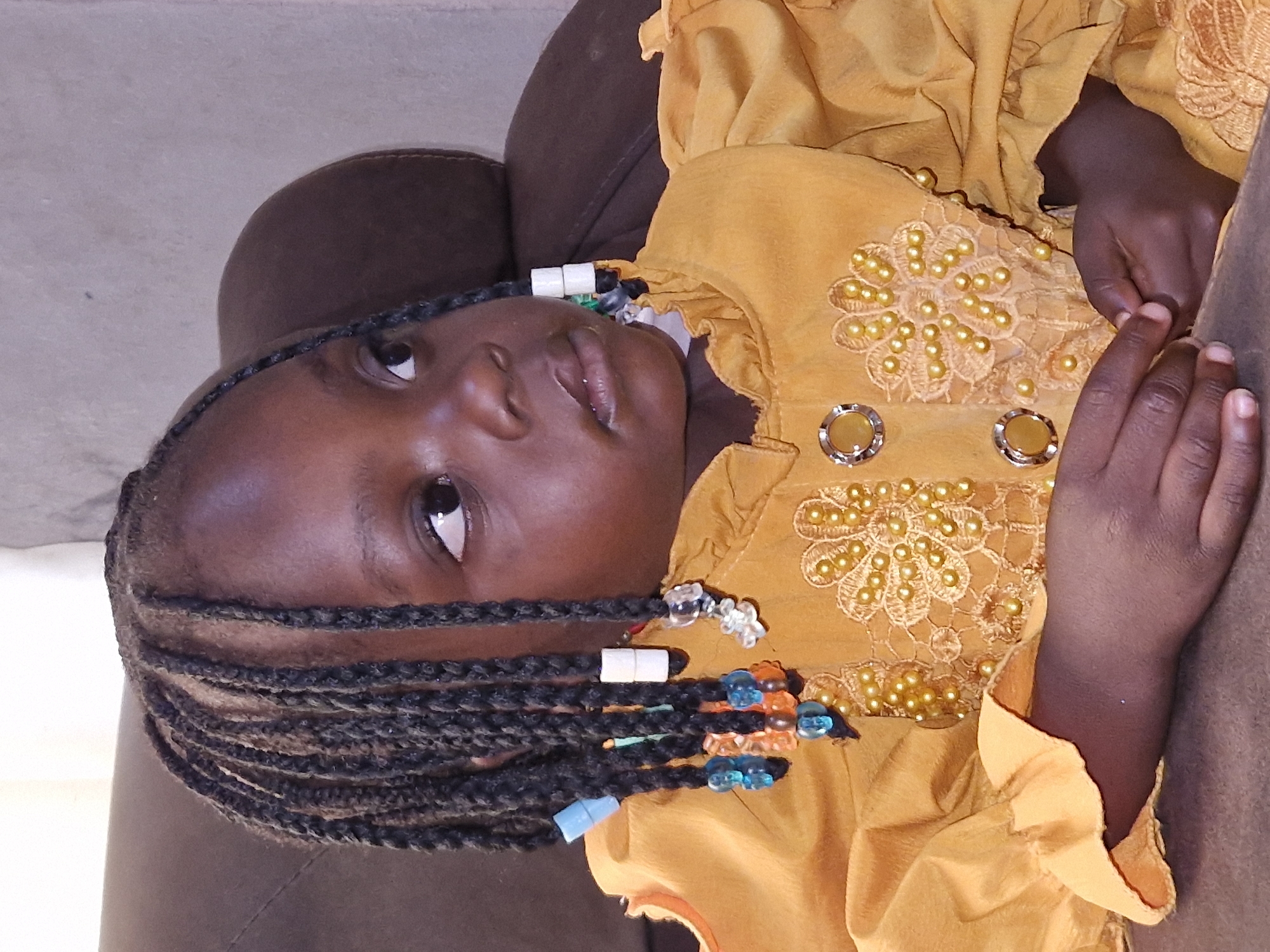 In the city of Jos, Nigeria, a young girl with braided hair adorned with beads is seated on a brown sofa. She is wearing a yellow dress with ruffled sleeves and decorative gold embroidery and beads on the chest. Her hands are clasped in her lap, and she is looking to the right, with a thoughtful expression. The background is blurry, showing the back of the sofa and a plain wall.