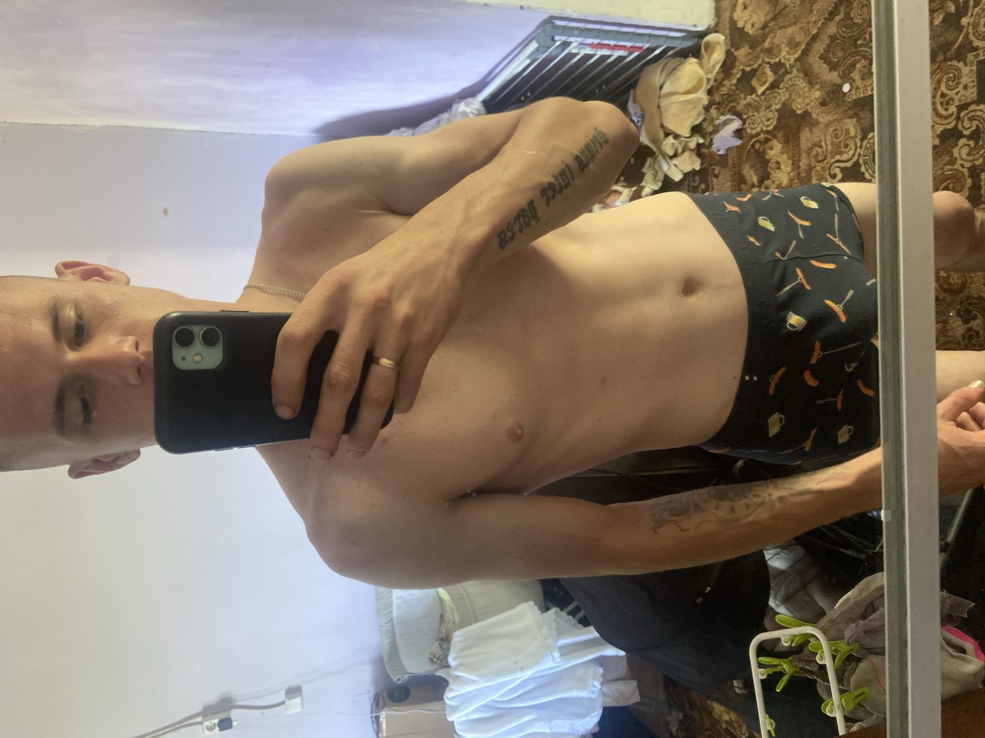 A man is taking a selfie in a mirror. He is shirtless, wearing patterned boxer briefs with images of beer mugs and hot dogs. He has a tattoo on his left arm and a ring on his right hand. The room appears to be a bedroom, with a patterned carpet and some laundry on the floor. A vent is visible on the wall behind him. The lighting suggests it is daytime. He is holding a smartphone in his right hand.