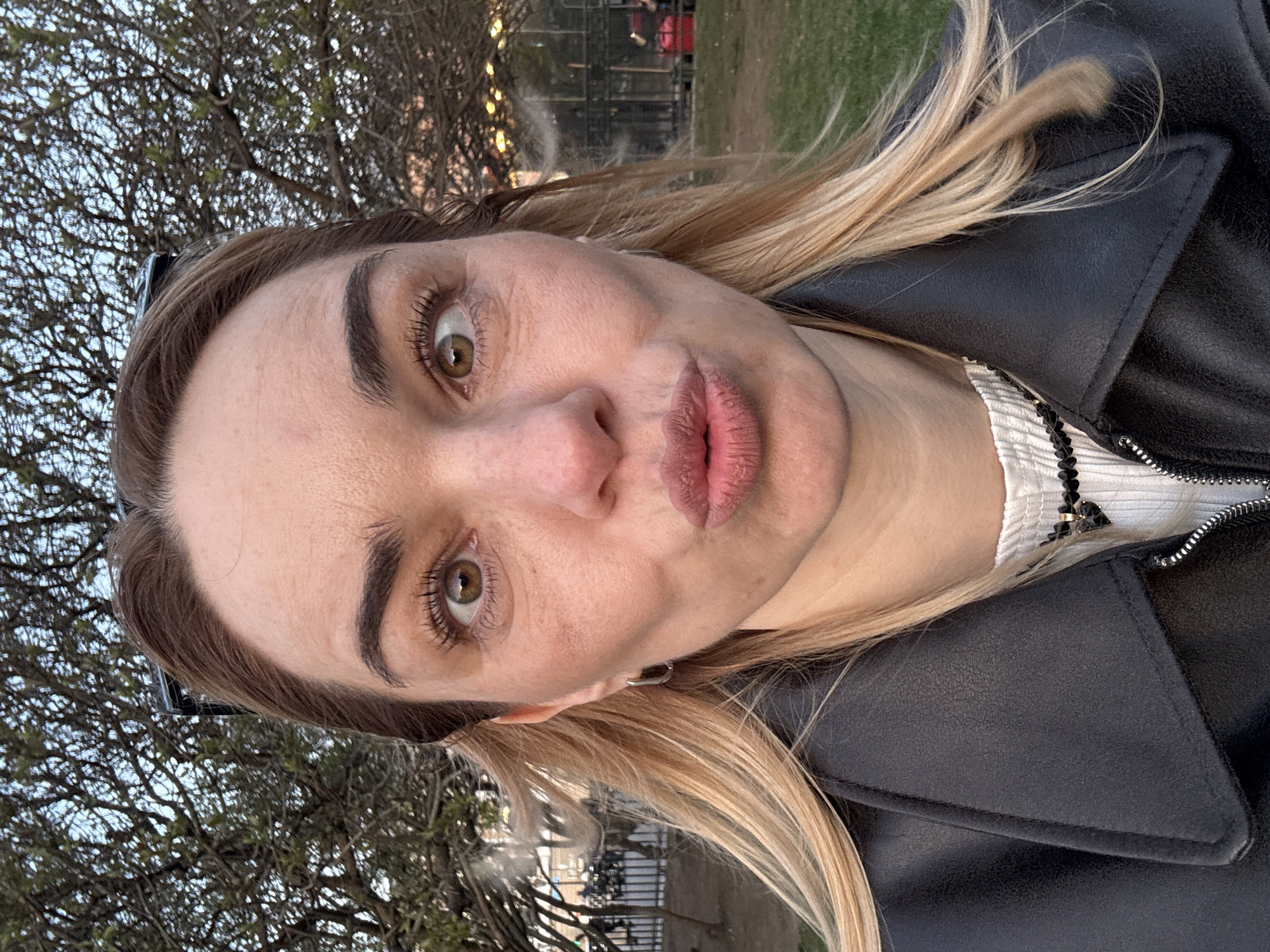 A fair-skinned female with blonde hair and hazel eyes is posing for a selfie outdoors. She wears a black leather jacket over a white top with black embellishments. The background shows trees and a fence, suggesting a park setting. The lighting indicates either early morning or late afternoon. The location could possibly be Saint Petersburg, Russia, but there is no way to prove it.