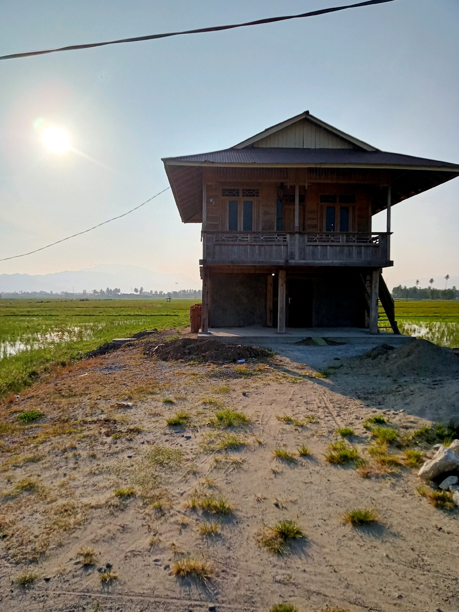 An elevated wooden house stands on stilts in a rural area of Pantungo, Indonesia, adjacent to a vibrant green rice paddy field. The house has a weathered wooden facade with visible grain, a dark corrugated metal roof, and a covered balcony area on the second floor. A wooden staircase leads to the entrance on the second floor. The ground floor appears to be unfinished concrete. In front of the house, a dirt path with sparse dry grass leads towards the viewer. The sun is low in the sky, casting a warm light and creating lens flare on the left side of the image. In the distance, a hazy mountain range is visible beyond the flat landscape of the rice paddies, which are partially flooded. Several palm trees dot the horizon. The overall scene depicts a serene, agricultural environment under a clear sky.