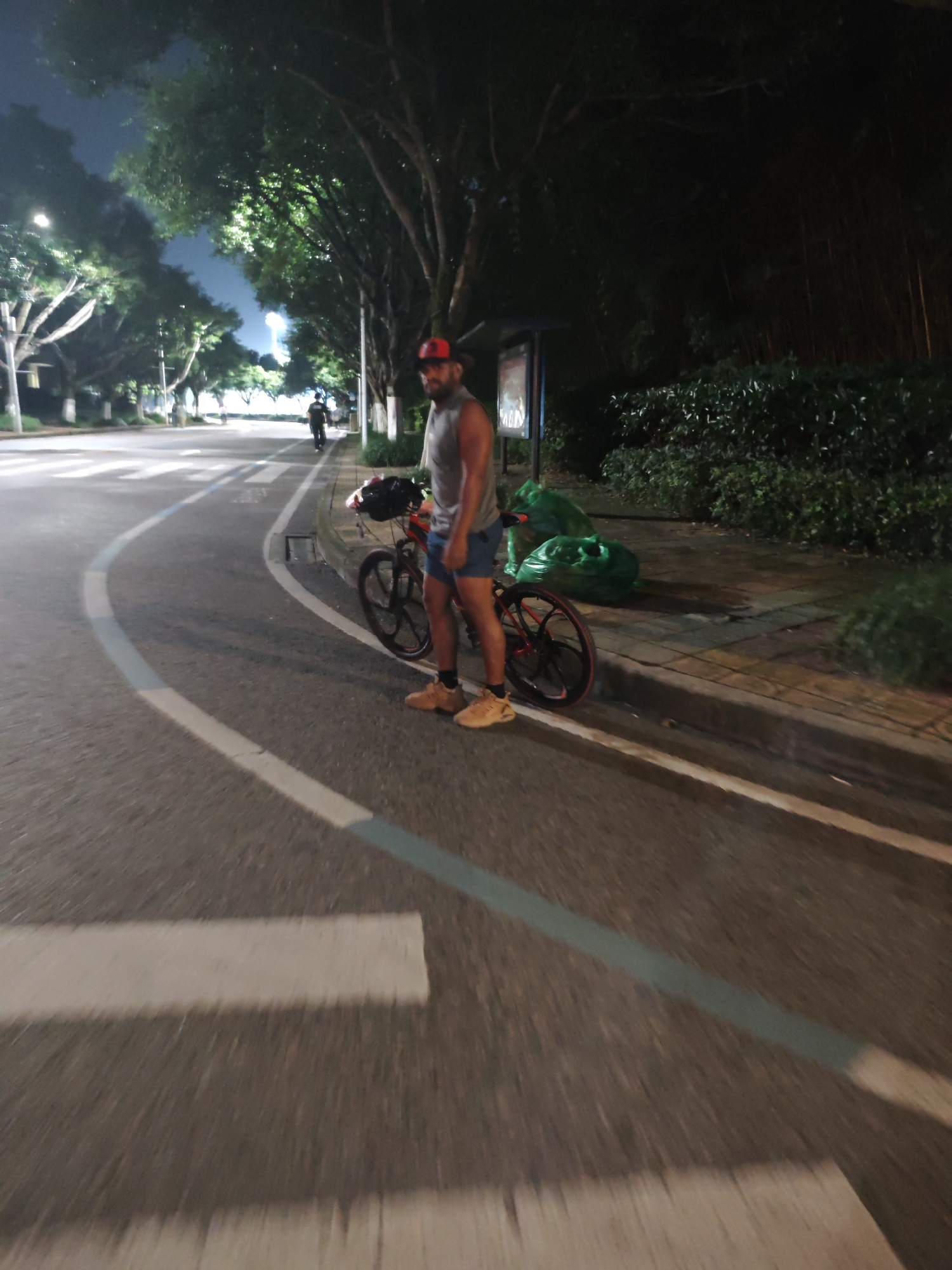 In the city of Chongqing High-tech Zone, China, at night, a man stands next to his bicycle on the side of a paved road. The man is wearing a grey sleeveless shirt, blue shorts, a red baseball cap, and tan sneakers. His bicycle is red with black rims. The road has white and light blue markings. In the background, there are trees with illuminated leaves, streetlights, and a few other people in the distance. Two large green bags are placed on the sidewalk behind the man.