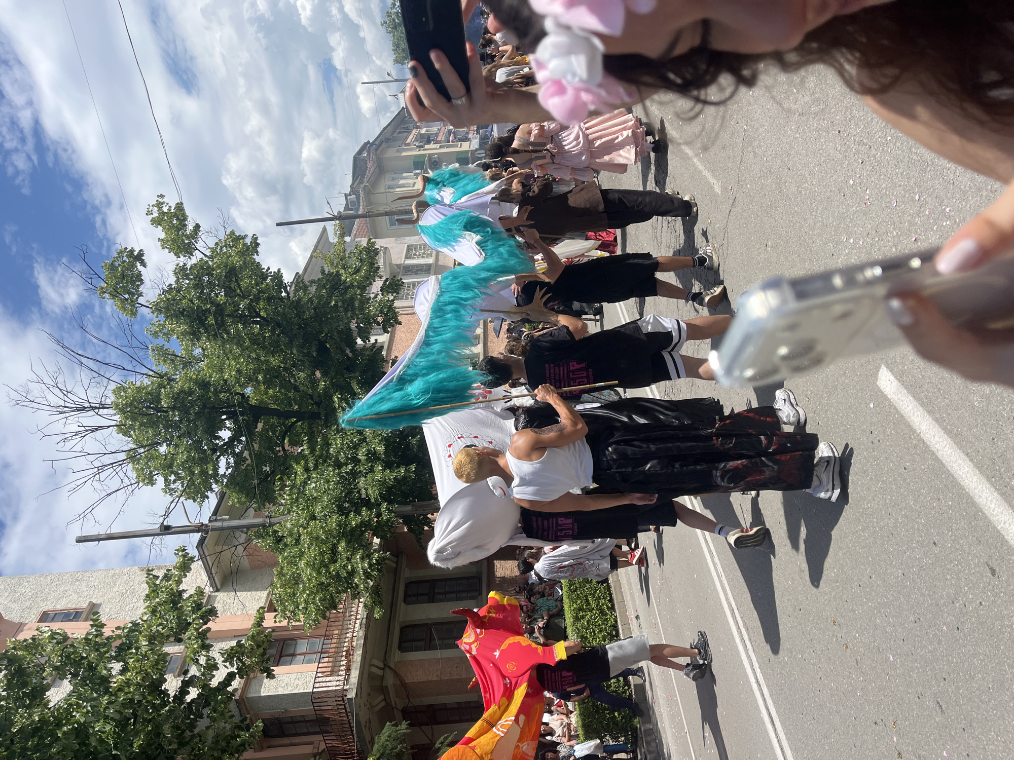 An outdoor parade or procession unfolds on a city street under a partly cloudy daytime sky in Unknown, Unknown. The street, paved with asphalt, features white double lines down the center and scattered pink petals.
In the immediate foreground on the right, a person with dark, curly hair and a pink floral headband (flower crown) holds a silver smartphone in a clear case, positioned for capturing the scene. Their lower right face, showing a slight smile, and manicured fingernails are visible.
The midground features numerous individuals participating in the procession, moving from right to left. A person with bleached blonde hair, wearing a white tank top and shiny black pants, holds a tall pole adorned with vibrant teal-turquoise feathers. Adjacent to them, another individual carries a large white banner or sheet, partially obscured, with a faint red circular design. To their left, a person wearing a black t-shirt with "5OP" in white lettering and grey shorts carries a large, red and orange bull-shaped figure on their head and shoulders. Further back, another participant holds a similar teal-turquoise feathered prop, which incorporates two white, horn-like elements. Various other participants are visible, including one in a long pink dress, walking with their back to the camera, and others in casual attire like t-shirts, shorts, and sneakers.
On the left side of the street, behind a low green hedge, a crowd of spectators observes the procession.
The background reveals multi-story buildings lining the street. The building on the left features a textured light-colored facade with peach-pink architectural accents, multiple windows, and balconies with dark metal railings. Buildings on the right are also multi-story, exhibiting various styles. Abundant green trees with dense foliage are interspersed, some partially obscuring buildings, while a tall, dark utility pole stands prominently in the upper mid-ground with overhead wires visible against the sky. Shadows are pronounced, indicating strong overhead sunlight.