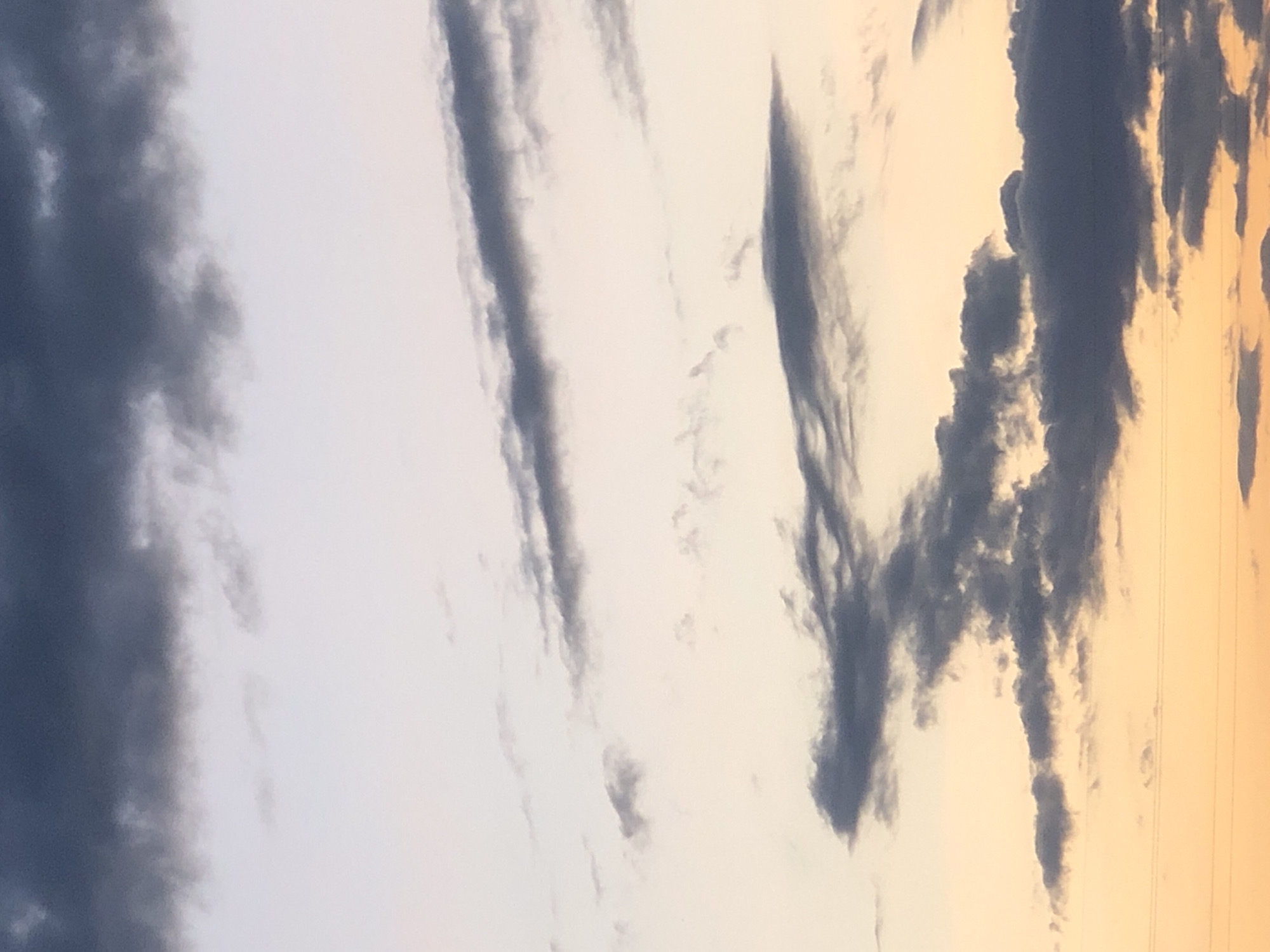 The image shows a sky filled with elongated, dark clouds. The color of the sky varies from light gray to orange near the horizon. Thin, straight lines suggesting electrical wires can be seen at the right edge of the image. The specific location in Krasnoyarsk, Russia is not discernible from the image.