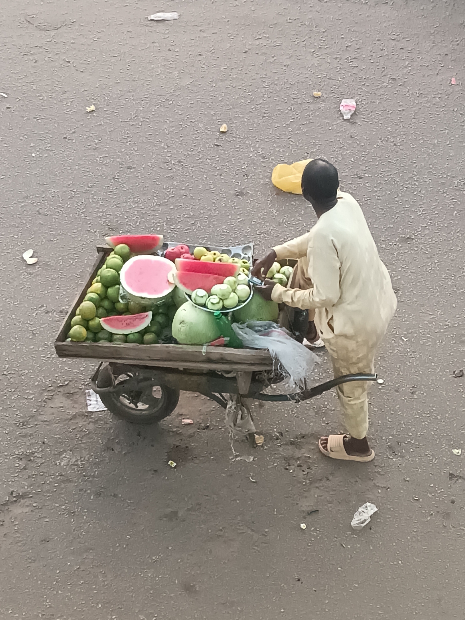 An elevated view captures a street vendor with his fruit cart in Bukuru, Nigeria. The central subject is a dark-skinned man, seen from behind, wearing a light-colored tunic and trousers with light-brown slip-on sandals. He stands next to a wooden, two-wheeled handcart laden with fresh produce.
The cart holds a variety of fruits: several large green watermelons, some of which are cut open to reveal vibrant pink flesh. A prominent pile of green, spherical fruits, likely limes or green oranges, occupies a significant portion of the cart. Smaller clusters of light green, round fruits, possibly guavas, are also arranged in bowls or containers on the cart.
The man appears to be tending to or arranging the fruits on his cart, with his hands near the smaller green fruits. The setting is an outdoor, unpaved or poorly paved area, possibly a street or market, characterized by a greyish, textured ground with scattered small debris and litter, including discarded plastic. The lighting suggests it is daytime. There is no clearly visible text.