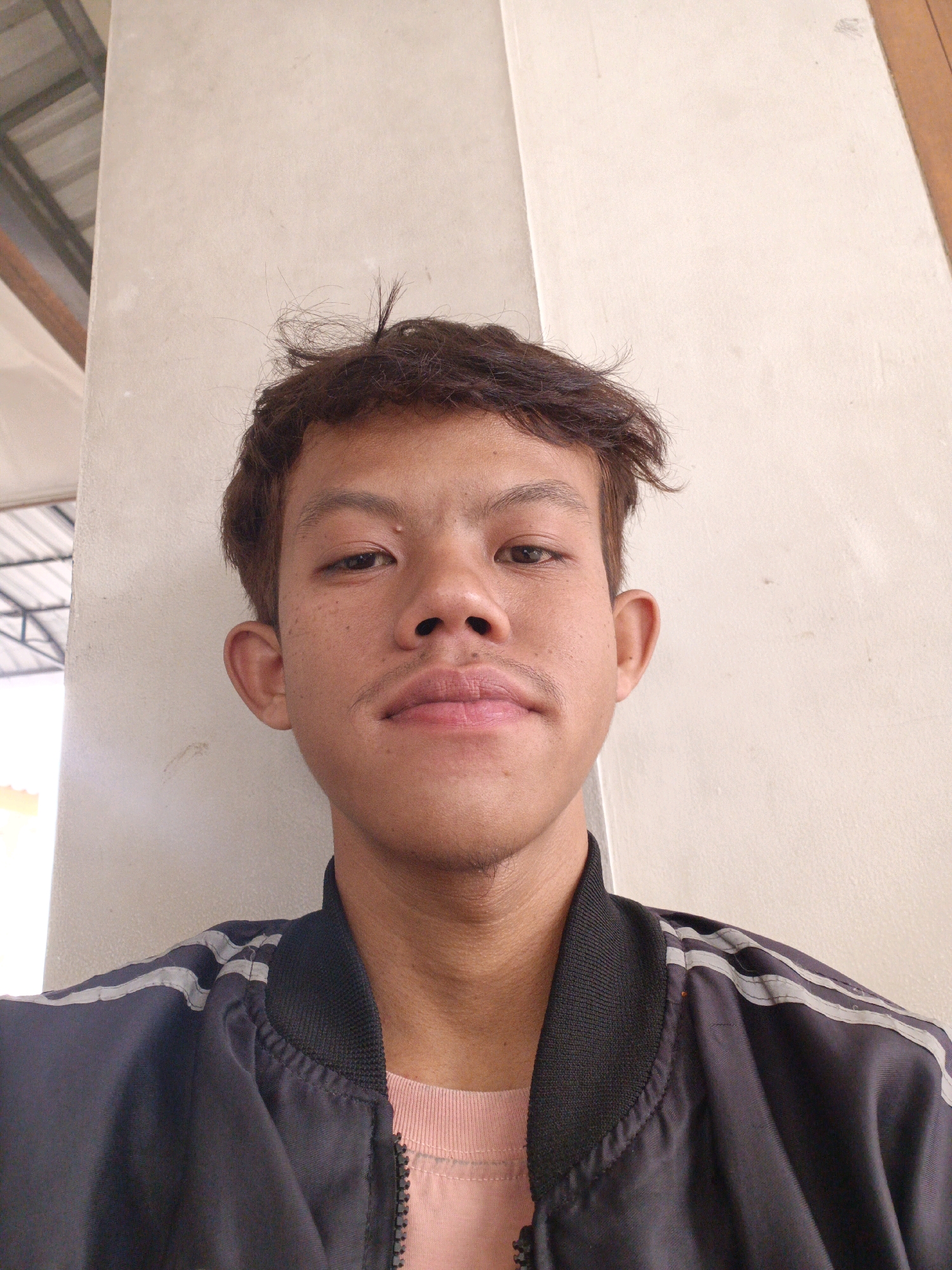 A young man with dark, messy hair and a light complexion is pictured from the chest up, leaning against a cream-colored wall. He has dark eyebrows, brown eyes, and a faint mustache. He is wearing a black jacket with white stripes on the shoulders and a pink t-shirt underneath. The background consists of a plain wall and a glimpse of a metal roof structure on the left. The image appears to have been taken outdoors, possibly in Kebonagung, Indonesia.