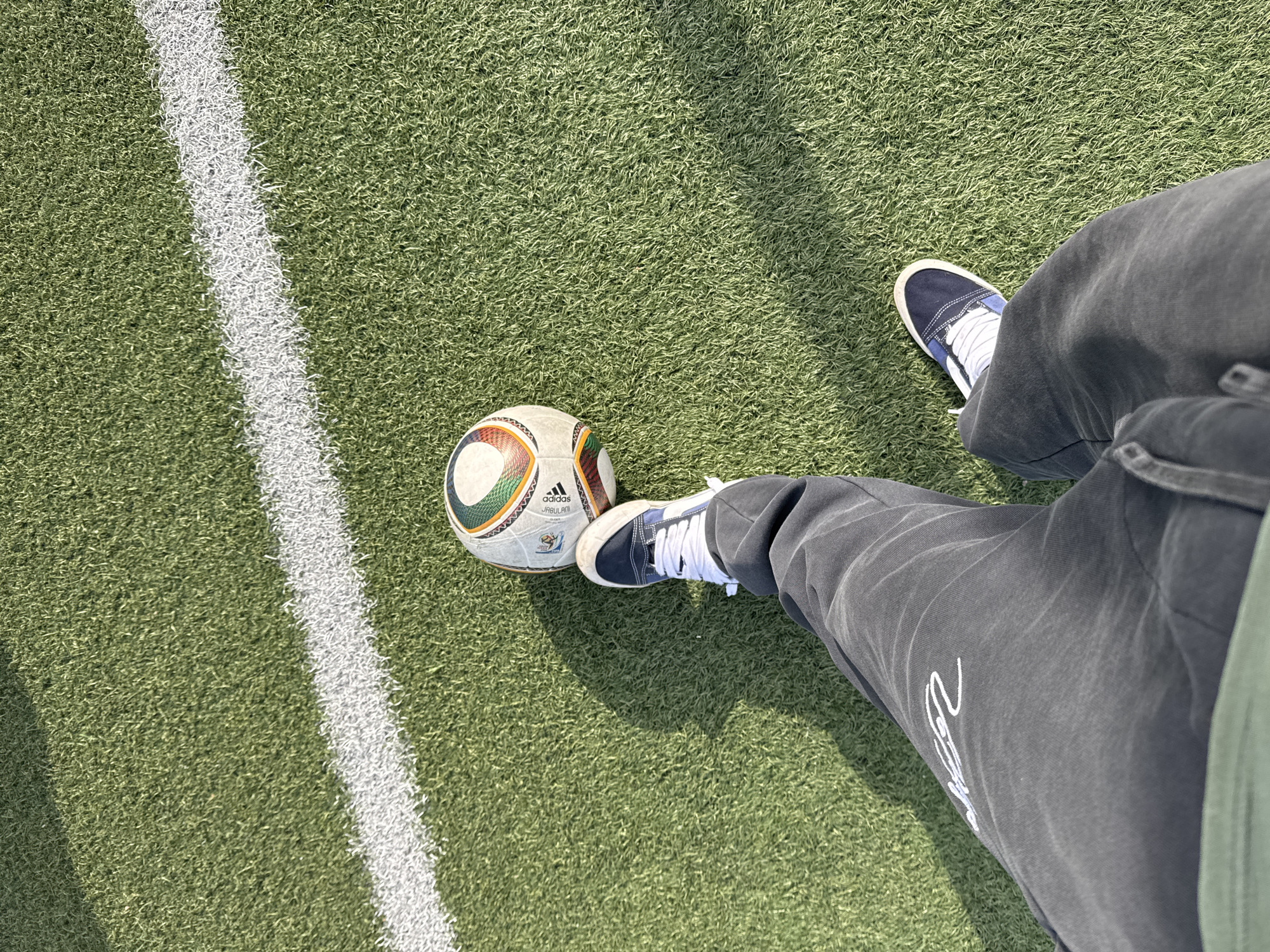 This high-angle, downward shot captures a person standing on an artificial turf soccer field in Sosnovo, Russia. The scene features the lower half of the person's body, including their legs and feet, positioned on the green pitch alongside a soccer ball and a prominent white field line.
The person is wearing dark gray, possibly sweatpants, with a white, stylized graphic or text visible on the left pant leg. They have blue and white lace-up sneakers on both feet. A classic Adidas Jabulani soccer ball, clearly marked with "Adidas" and "JABULANI" text, lies on the turf near their left foot. The ball features its distinctive white panels with colorful, intricate patterns.
The strong shadows cast by the person and the ball indicate bright, direct sunlight, suggesting a daytime setting. The overall impression is of someone on a soccer field, perhaps about to kick the ball or taking a break from playing.