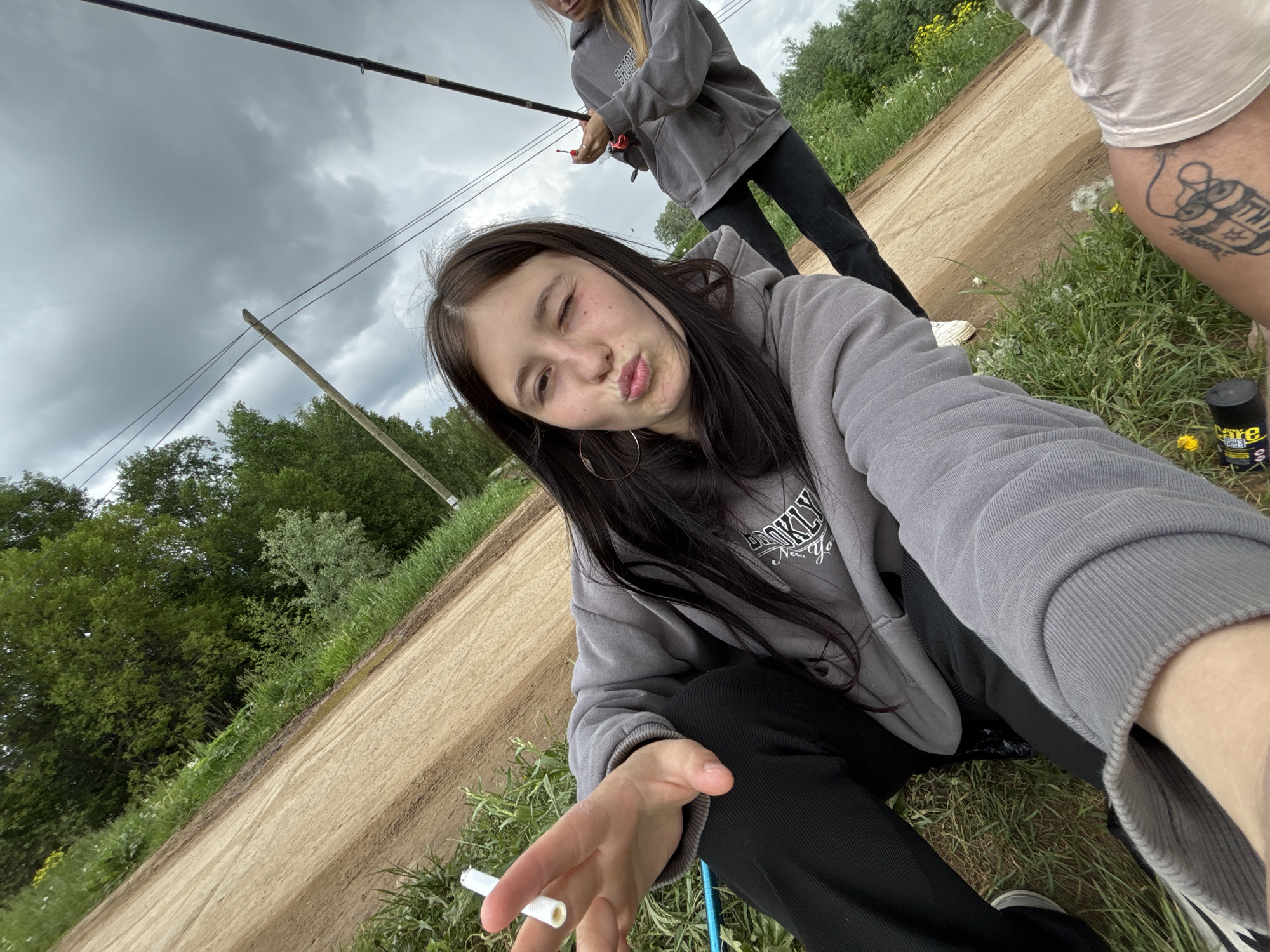 The image depicts a young woman taking a selfie outdoors. She is winking and making a kissing face at the camera. She's wearing a grey hoodie with the words "Brooklyn New York" on it and black pants. She is holding what appears to be a cigarette in her hand. Another person is visible in the background, also wearing a grey hoodie, and holding a fishing rod. A third person is partially visible on the right side of the frame, with a tattoo on their leg. The setting appears to be rural, with a dirt road and grassy areas. There are trees in the background, and the sky is overcast with dark clouds. Based on the location, the scene might be set in the vicinity of Фоки, Russia, given the user query.