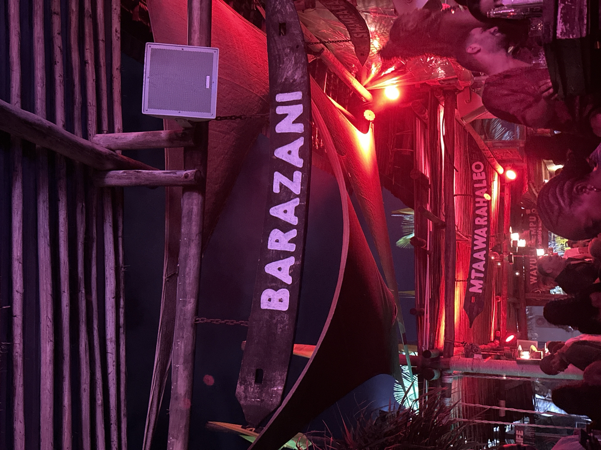 The image shows an indoor scene in Unknown, Unknown, with a tiki bar-like aesthetic. The decor includes bamboo-style vertical slats, wooden beams, and a stylized boat sail bearing the word "BARAZANI". A square, white speaker is mounted on a post near the sail. Red lighting permeates the scene. People are present, some partially visible in the background, interacting near the bar. The word "MTAAWARAHALEO" is visible on a vertical banner. There are also objects on the bar surface, possibly glasses or decorative items, reflecting in the surface. The overall impression is of a dimly lit, lively bar environment.