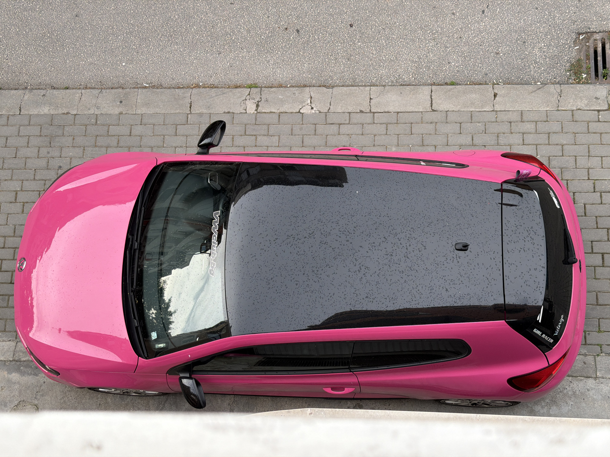 An overhead shot shows a bright pink, two-door vehicle with a black roof parked on a cobblestone street adjacent to an asphalt road. The car is positioned vertically in the frame, with its front facing downwards. Raindrops are visible on the car's black roof, indicating recent precipitation. A sticker reading "GIRL RACER Victoriya" is affixed to the rear of the car. The environment suggests an urban setting in Plovdiv, Bulgaria, with the cobblestone pavement and the texture of the asphalt road being characteristic of the city's older districts or pedestrian areas. A white wall is visible on the left side of the frame, bordering the car. A storm drain grate is present on the right side of the asphalt road. There are no people visible in the image.