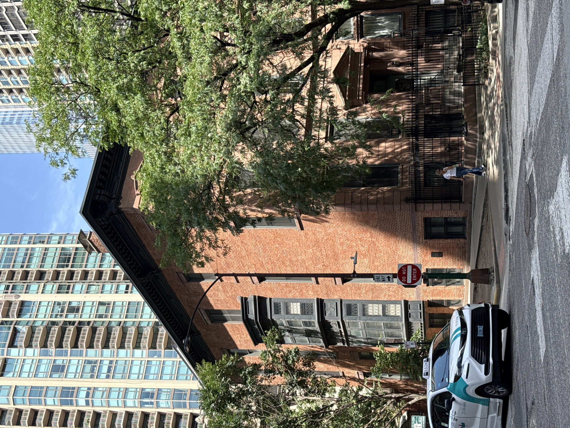 This is an outdoor, eye-level, rotated shot of a street scene in Chicago, USA. The sun is shining, casting dappled shadows on the street and buildings, suggesting it is daytime.
The foreground features a white and teal-colored taxi cab with the number 5378 prominently displayed on its side. The taxi is parked on the right side of the street, facing away from the viewer. Next to the taxi, a "DO NOT ENTER" sign is visible, mounted on a pole with a "ONE WAY" sign.
A person in a white shirt and blue jeans is walking on the sidewalk to the left of the "DO NOT ENTER" sign. Behind the person, a brick building with dark-trimmed windows and ornate architectural details stands tall. A black metal fence runs along the front of the building.
In the background, a modern high-rise building with a grid of blue windows and balconies dominates the left side of the frame. A large, leafy green tree is situated between the brick building and the high-rise, its branches extending over the brick building. The overall scene depicts an urban environment where older architecture coexists with contemporary structures.