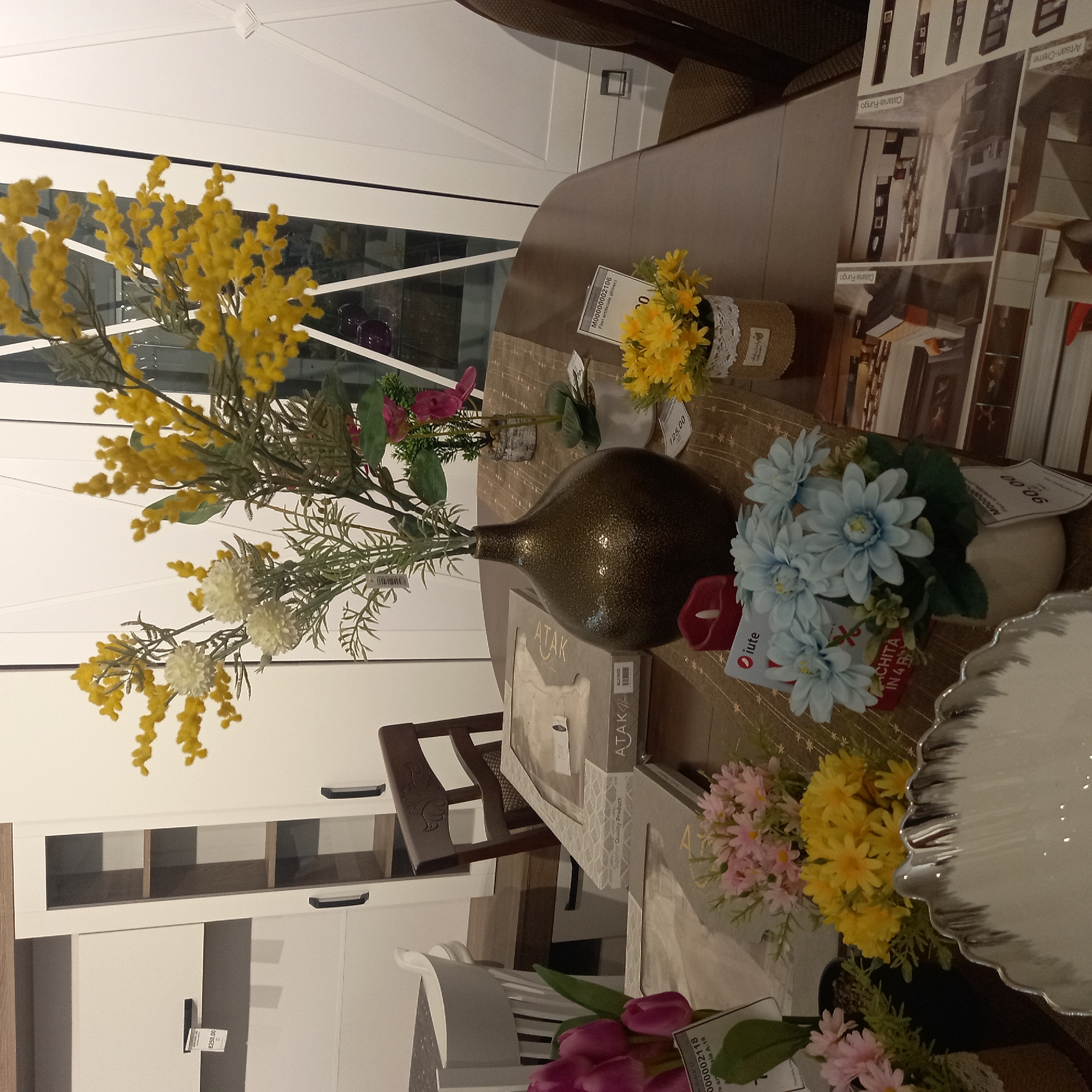 This media file depicts a collection of artificial flowers and decorative items arranged on a tabletop, likely in a retail or home setting. The subjects include several bouquets of artificial flowers in various colors, predominantly yellow, white, and pink, with some blue flowers also visible. These are placed in simple vases or containers. A large, dark, metallic vase is a central object, holding a prominent yellow floral arrangement.
Other objects on the table include decorative books or catalogs with visible text such as "ATAK" and "Catania-Fungo." Price tags with numbers like "125.00" and "8250.00" are also present, suggesting a retail environment. A red decorative candle is positioned near the blue flowers. The overall scene is one of tasteful arrangement and display of decorative elements.
The setting appears to be indoors, with a suggestion of modern furniture, possibly a dining table or display table. The lighting is bright, indicating daytime. There are no people visible in the media file, and no specific activity or event is taking place. The visible text provides product names and pricing information. The background shows hints of furniture and white walls, possibly a showroom or a well-decorated room. The image is oriented such that the objects appear to be viewed from a slightly elevated perspective. The presence of catalog images related to furniture in the background further supports the idea of a retail or interior design context.