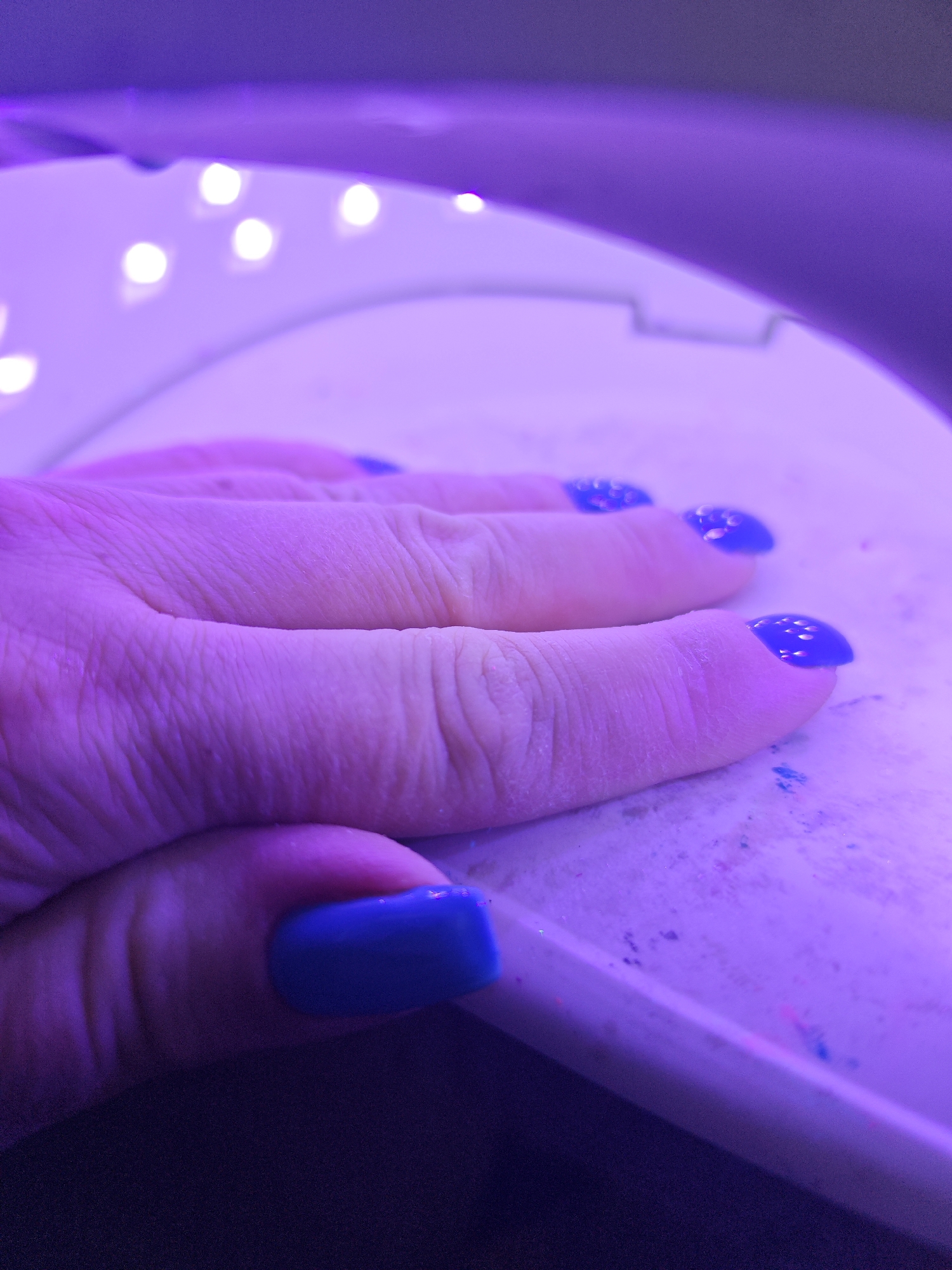 A close-up shot depicts a hand with manicured fingernails coated in glossy blue polish positioned under a UV nail lamp, likely during a gel manicure session. The hand is resting on a surface, presumably the base of the lamp or a nearby table. The lamp, visible above the hand, has a series of white LED lights. The lighting casts a strong purple hue over the scene, affecting the colors of the hand and surrounding surfaces. The image does not provide any information about the location of Марковский, Russia. The image primarily focuses on the hand and nail lamp.