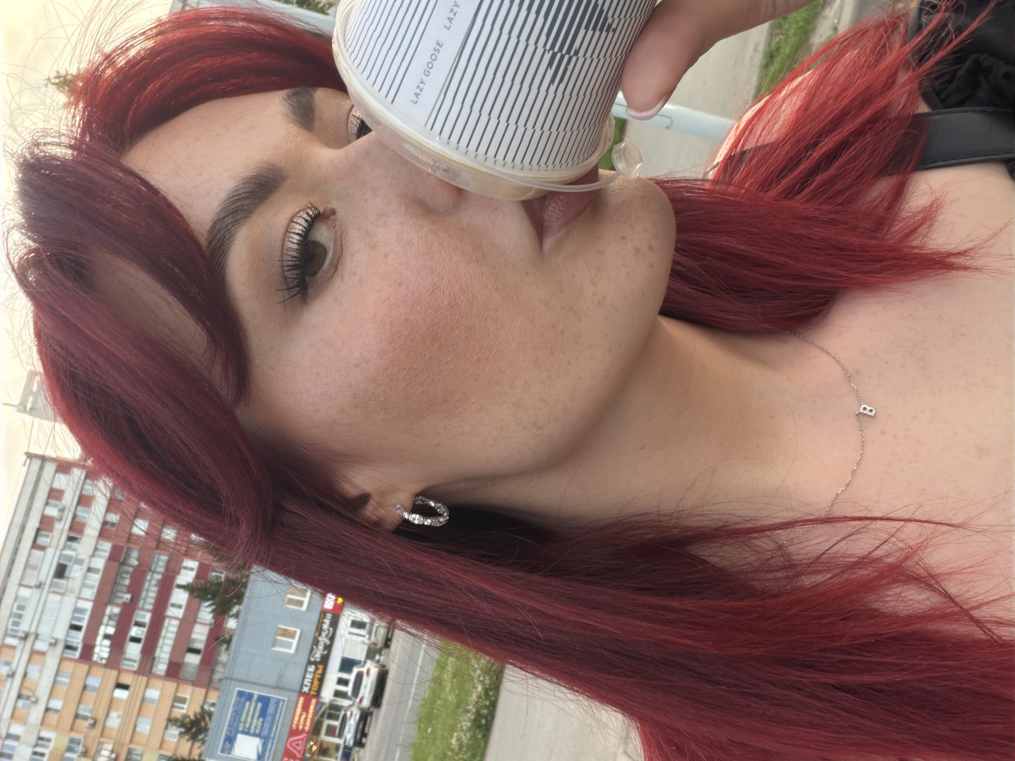 A close-up shot shows a young woman with bright red hair, freckles, and dramatic makeup. She is holding a white paper cup with a black and white striped design and drinking from it. She wears a silver hoop earring in her left ear and a delicate silver necklace with a pendant that appears to be the letter "B". In the background, there are tall apartment buildings and trees, suggesting an urban setting. The presence of buildings similar to those found in Tolyatti, Russia, along with the context of the image being a public street scene, implies it might be located in that city. The overall impression is a casual, outdoor moment captured on a bright day.
