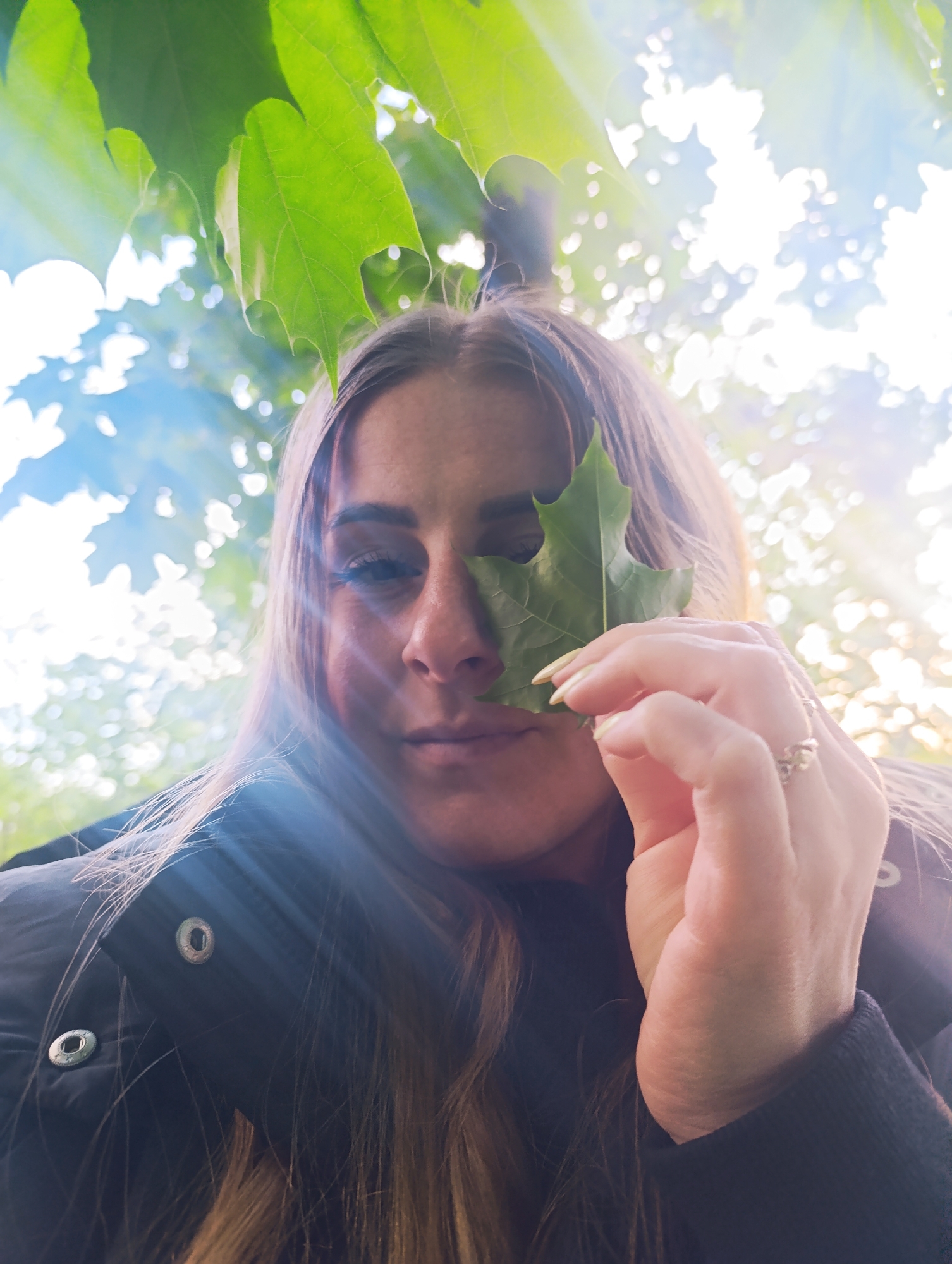 A woman holds a green leaf in front of her face, partially obscuring her nose and right eye. She has long blonde hair and is wearing a black jacket. Above her are more green leaves and a glimpse of blue sky with bright sunlight shining through. The location is unknown.