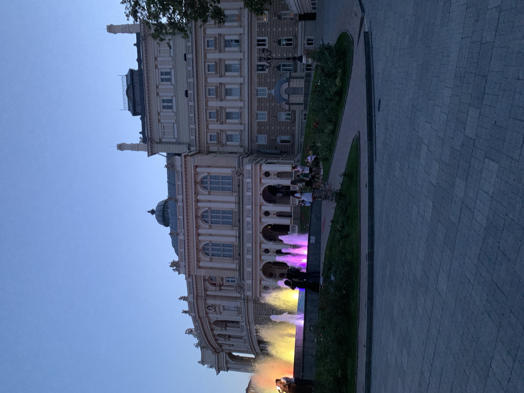 The image captures the Odessa Opera and Ballet Theater in Odesa, Ukraine, at dusk. A colorful fountain with illuminated jets of water in yellow, pink, and purple is in the foreground. Several people are gathered around the fountain, some standing and some sitting. The grand architectural facade of the opera house, with its multiple arched windows and ornate detailing, serves as the backdrop. The sky is a clear, fading blue. Paved walkways and manicured greenery surround the fountain and the building.