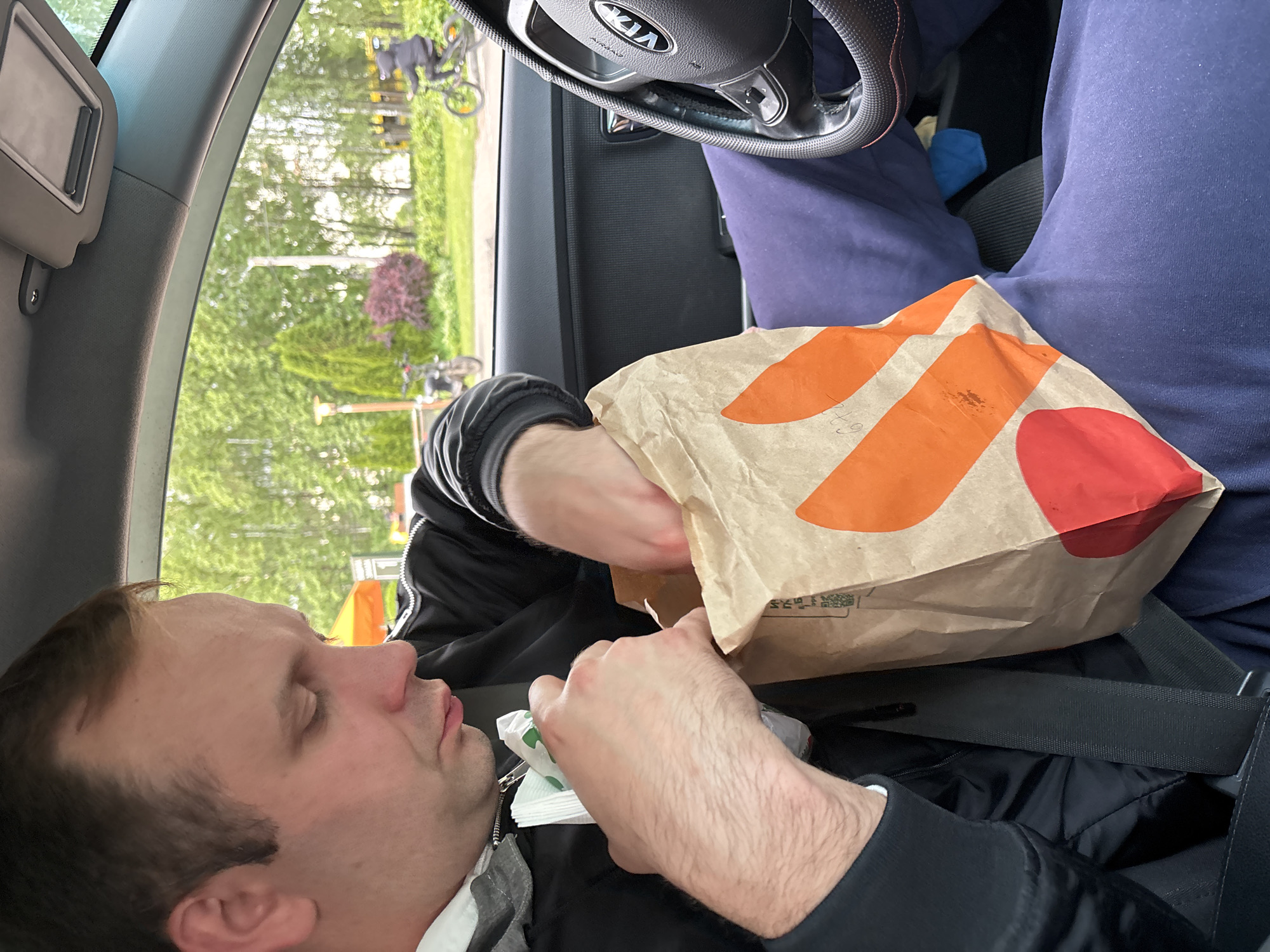 A man is seated in the driver's seat of a Kia vehicle, with the steering wheel visible in the background. He is wearing a black jacket and is holding a Taco Bell paper bag. He appears to be rummaging through the bag, possibly retrieving food or napkins. He has brown hair and is wearing a seatbelt. Through the car window, trees and a bicycle rack are visible, suggesting an outdoor setting, possibly in Rusanovo, Russia, given the location instructions.