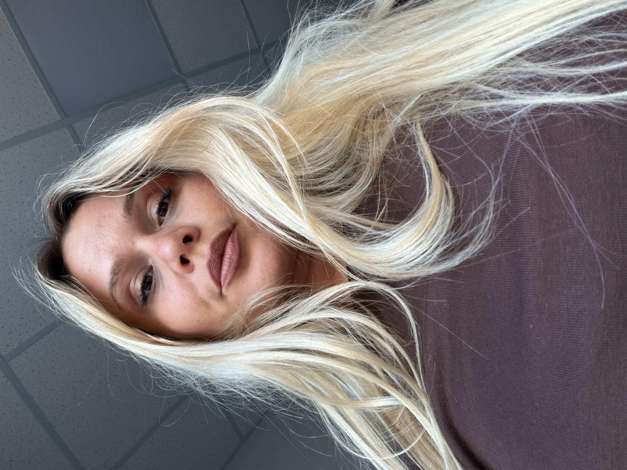 A selfie shows a woman with blonde hair, brown eyes, and light-colored lips. She wears a brown top. The background appears to be a ceiling with a grid pattern. There is no indication of location in Sarapul, Russia.