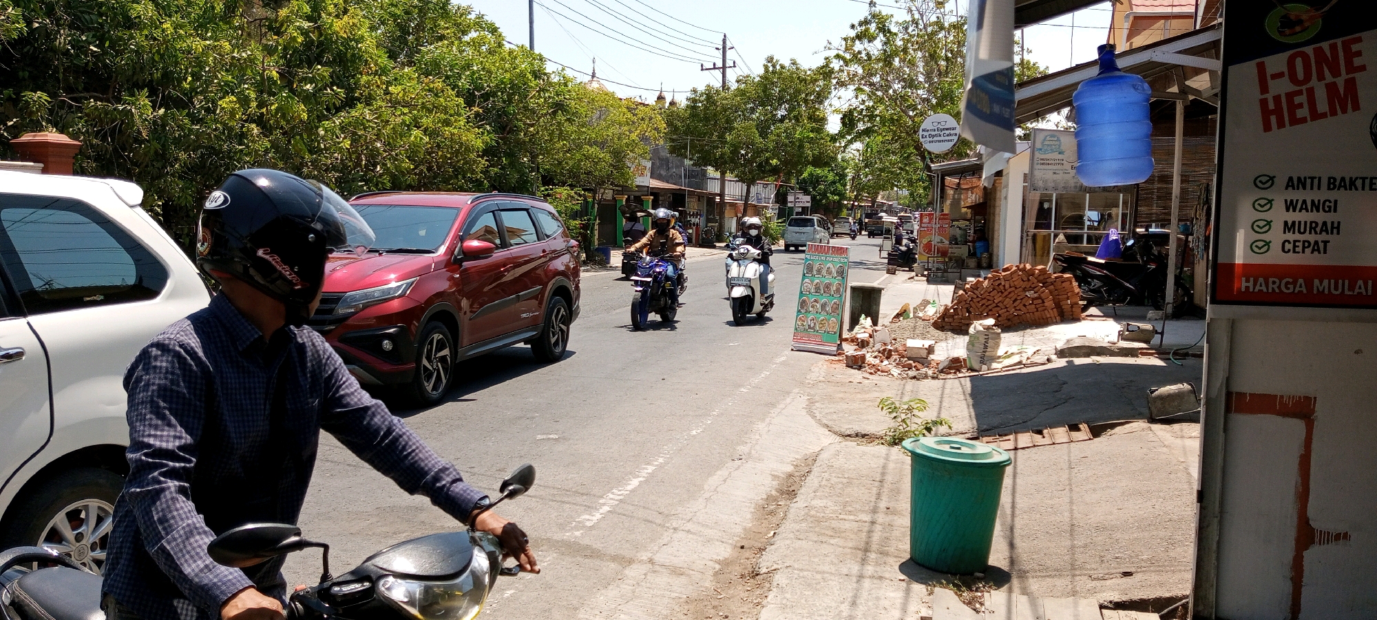 The image depicts a street scene in Sengkang, Indonesia, on a bright, sunny day. In the foreground, a person wearing a black helmet and a dark, checkered long-sleeved shirt is seated on a motorcycle, holding the handlebars. To the left, the rear portion of a white car is visible. Further down the street, a red SUV is driving away from the camera. Several other motorcycles with riders are also on the road, moving in the same direction. The street is lined with buildings on the right side, one of which features a sign with the text "I-ONE HELM" and bullet points listing "ANTI BAKTERI," "WANGI," "MURAH," and "CEPAT." A large blue water jug is suspended from an awning near this building. Construction materials, including stacks of bricks and bags of cement, are piled on the sidewalk. The environment includes mature trees and overhead power lines, suggesting an urban or semi-urban setting.