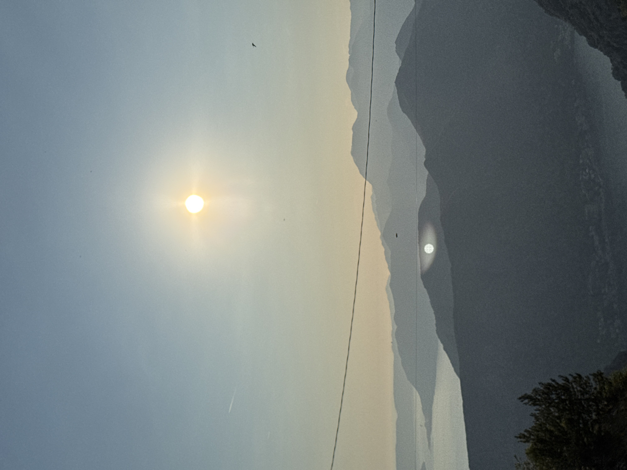 The image depicts a mountainous landscape at what appears to be sunrise or sunset. The sun, a bright, luminous orb with a halo effect, is positioned in the upper center of the frame, casting a warm glow. Multiple layers of silhouetted mountains recede into the distance, their forms softened by atmospheric haze. A body of water, likely a lake or fjord, is visible in the middle ground, reflecting the sky. A thin, dark power line stretches horizontally across the mid-section of the image, bisecting the view of the mountains. In the foreground, the dark silhouette of dense foliage, possibly trees, occupies the bottom left corner. The overall scene suggests a tranquil and natural environment, with hints of human presence indicated by the power line. The image has a slightly grainy texture, characteristic of older photographic processes or certain digital filters. The specific location in Žanjev Do, Montenegro, is not explicitly visible within the image itself, but the mountainous terrain and the presence of a large body of water are consistent with the geography of the region.