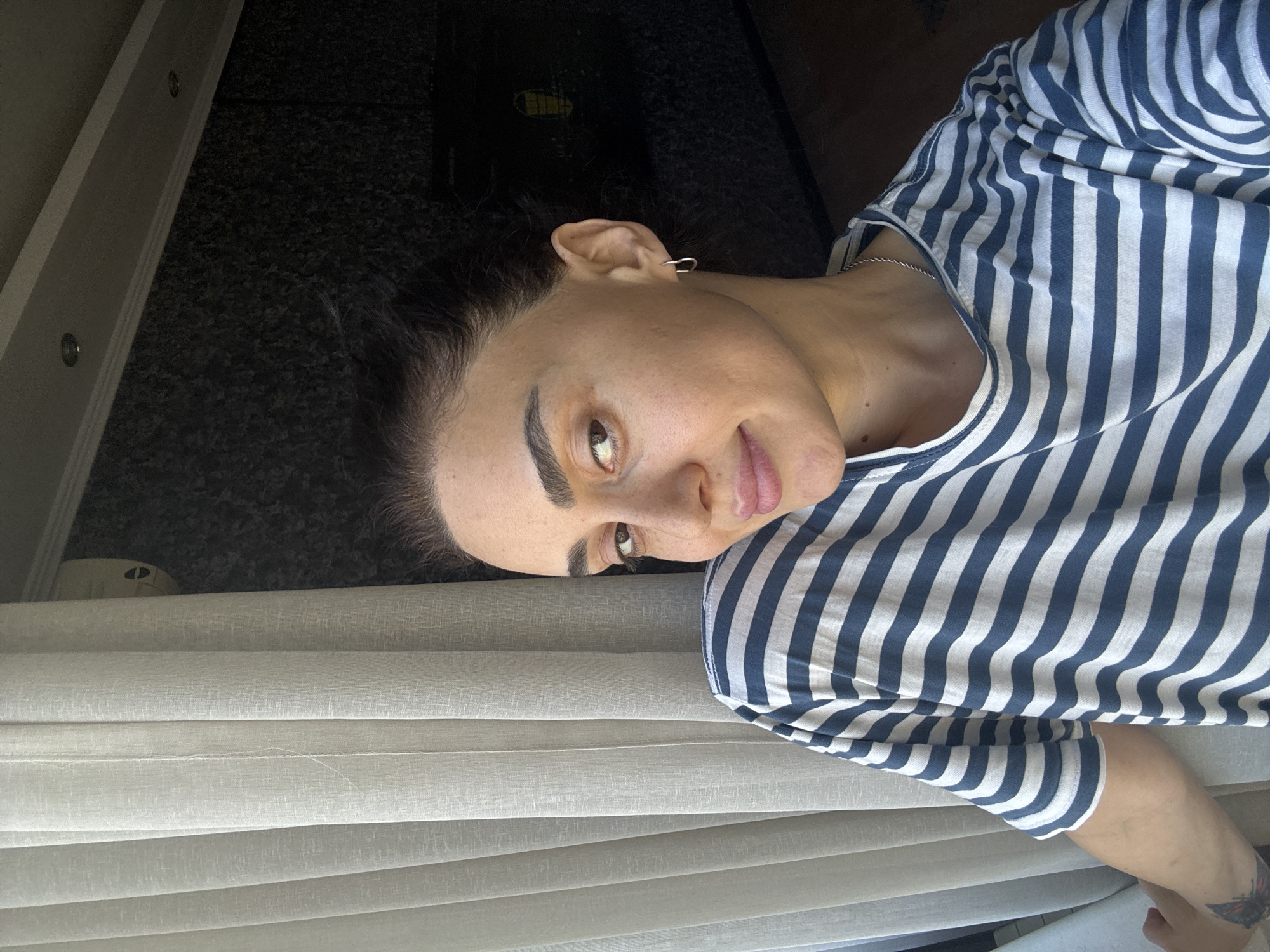 In an indoor setting with natural light filtering through a sheer white curtain on the left, a woman with dark hair pulled back and prominent eyebrows looks towards the camera. She is wearing a blue and white striped t-shirt. Her gaze is directed slightly to her right, and she has a slight smile. A small, heart-shaped earring is visible in her left ear. The background on the right consists of a dark, textured wall, possibly wallpaper, with some lighting fixtures visible on the ceiling above. The scene suggests a casual, personal moment captured at home, potentially in a residential area of Tbilisi, Georgia, though no specific landmarks or identifying features of the city are present.
