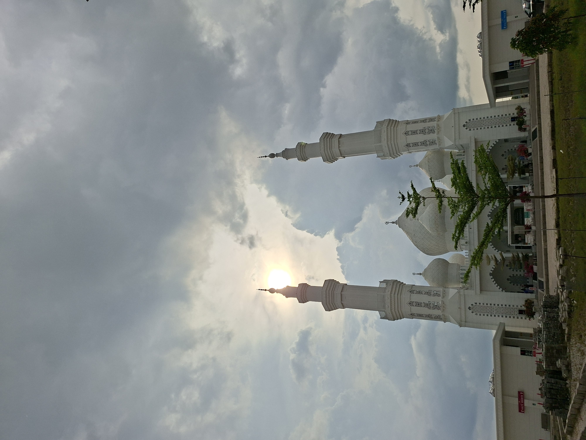 The image captures the Baiturrahman Islamic Boarding School Mosque in Meunasahaluedrienlhoksukon Barat, Indonesia. The mosque, predominantly white, features two prominent minarets and several domes. The sun is positioned behind one of the minarets, creating a backlit effect against a cloudy sky. The mosque is situated next to a building, with a green grassy area separating the two structures. Some trees are visible near the mosque. Construction materials and a sign with illegible writing are also present.