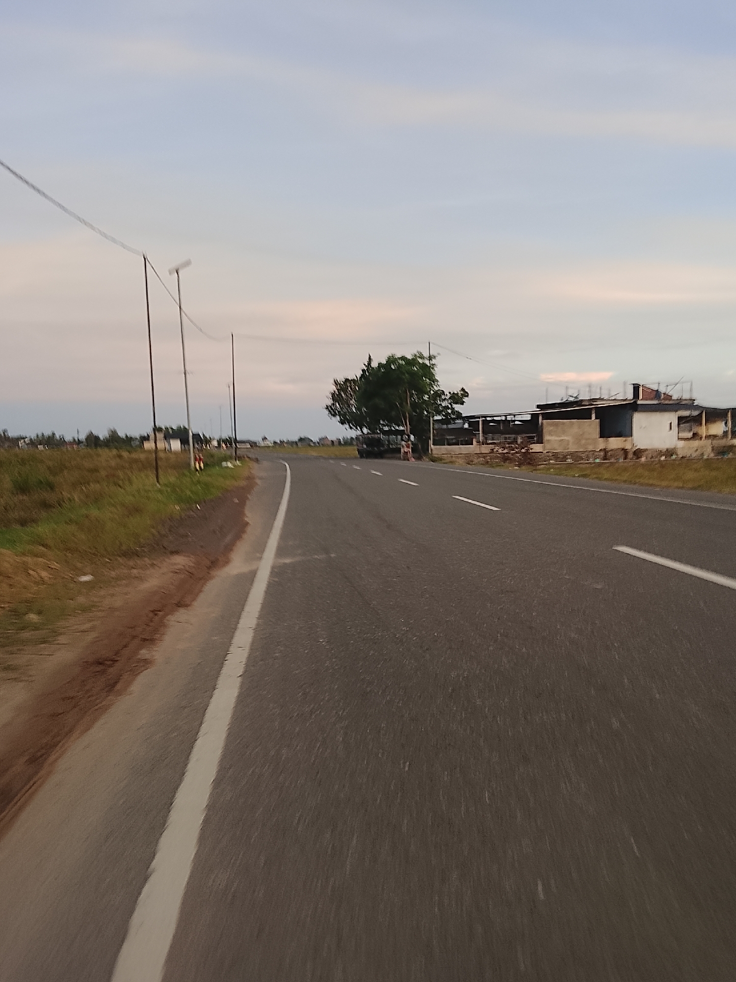 The image depicts a roadside scene in Dayah Kruet, Indonesia, under a pale, cloudy sky. The foreground is dominated by an asphalt road with a clear white lane marker running along the right side. The road curves gently to the right. To the left of the road, a grassy embankment with some reddish-brown soil is visible. Several utility poles with wires stretch across the scene. In the middle ground, a large tree stands near the roadside, and beyond it, several buildings are under construction or appear unfinished, with exposed concrete and some roofing materials. A dark-colored truck is parked near the buildings. The background shows more vegetation and a hazy horizon. The scene appears to be captured from a moving vehicle. There are no clearly identifiable people in the image.