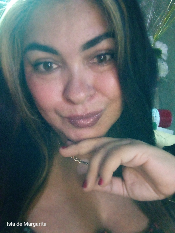 A woman with long dark hair and lighter streaks poses with her hand touching her chin. She has dark eyes, full lips with shimmery lipstick, and defined eyebrows. She is wearing a delicate silver chain necklace. In the background, there is greenery, a white flower, and what looks like tinsel. "Isla de Margarita" is printed at the bottom left corner of the image.