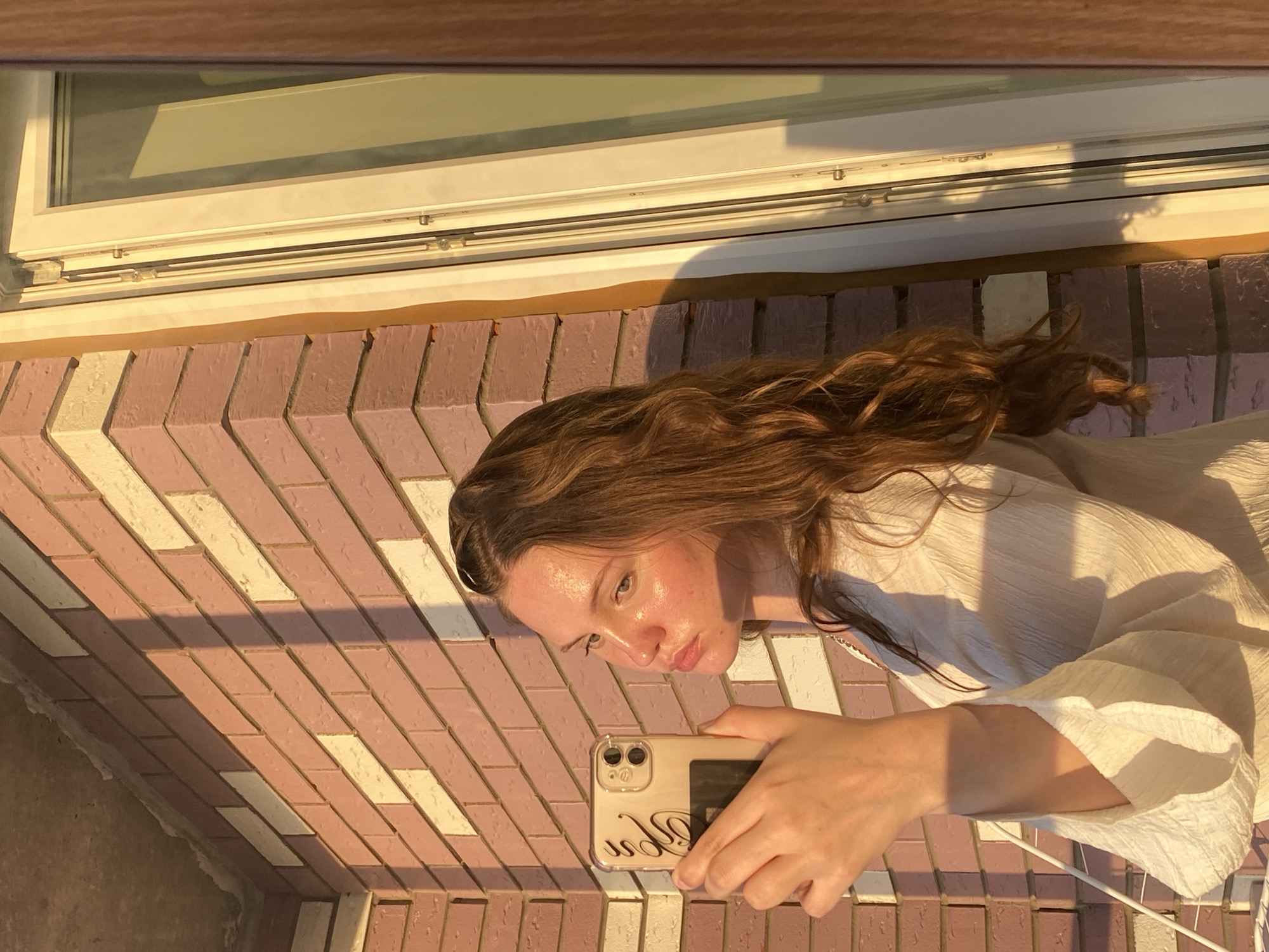 The image shows a young woman holding a smartphone to take a selfie. She has long, wavy brown hair and is wearing a light-colored, textured top. The background consists of a brick wall with alternating pink and cream-colored bricks, and a portion of a white window frame. The scene is illuminated by warm, golden hour sunlight, casting long shadows. The context of Izhevsk, Russia, is not visually discernible from the image itself.