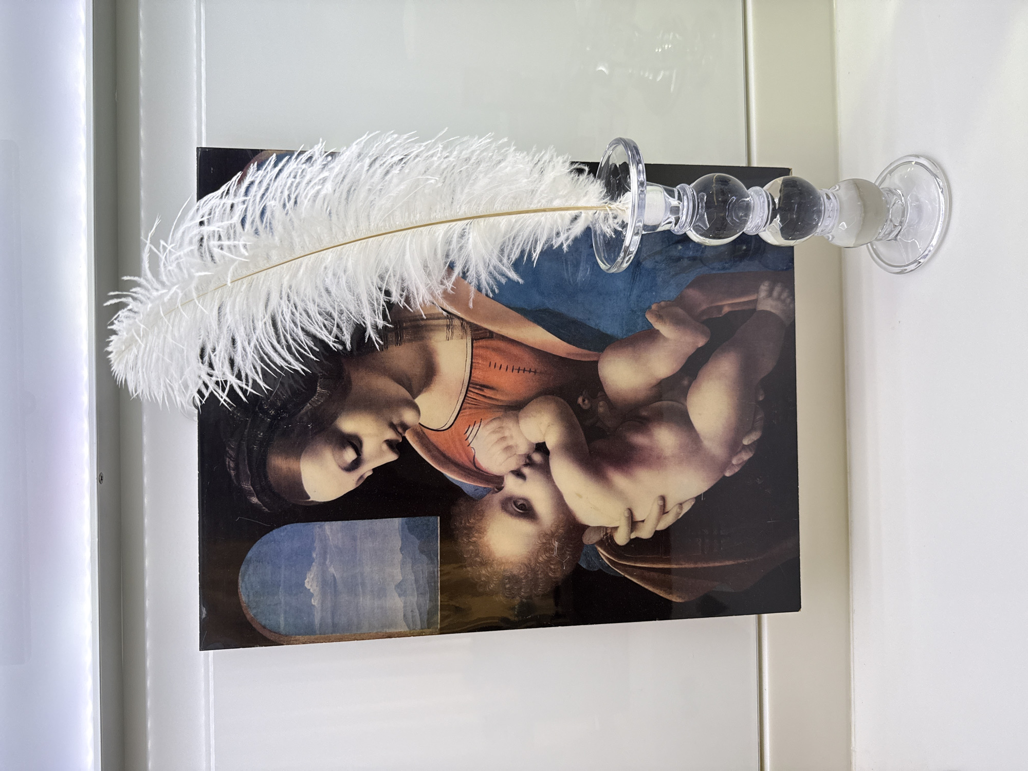 An image of the "Madonna Litta" painting, combined with a glass candlestick and a white feather. The painting is affixed to a white surface, possibly a wall or door. The candlestick, also affixed to the surface, lies horizontally across the painting and holds a white feather. There is no indication of the image's location in Votkinsk, Russia.