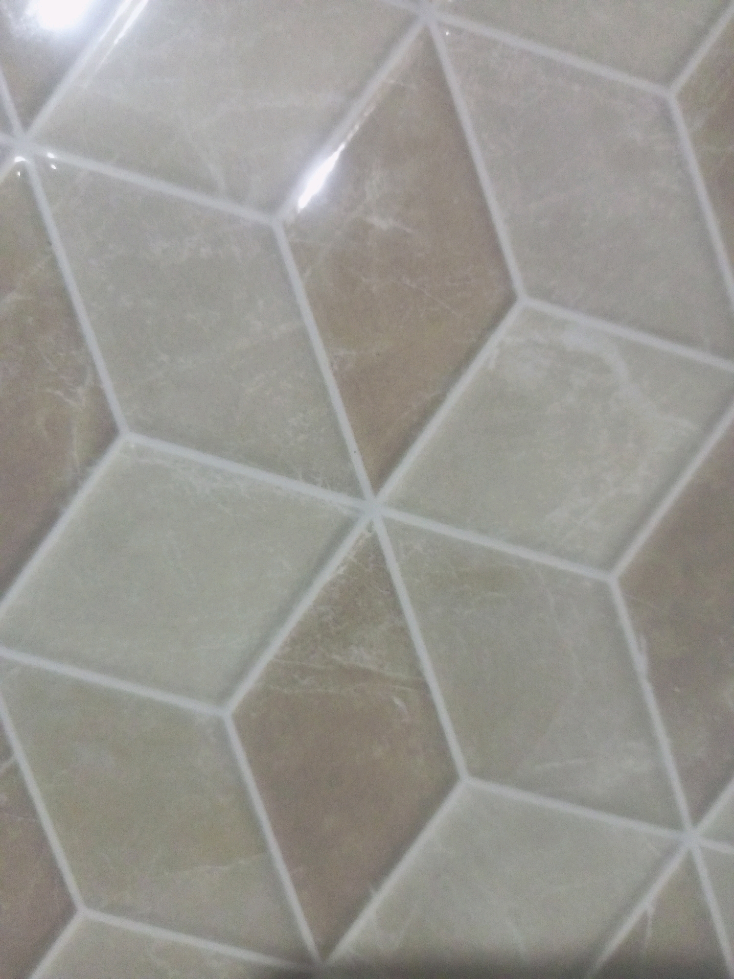 The image shows a close-up of tiled flooring with a geometric, three-dimensional pattern that creates the illusion of cubes. The tiles are a blend of light beige and taupe colors with subtle veining, resembling natural stone. The grout lines are a bright white, clearly defining the rhombus shapes that form the overall design. The lighting creates highlights and shadows, enhancing the perceived depth of the pattern. There are no people or any other objects visible in the image, and no specific location in Poka'an, Indonesia can be identified from the provided visual information. The scene is a static, indoor environment.