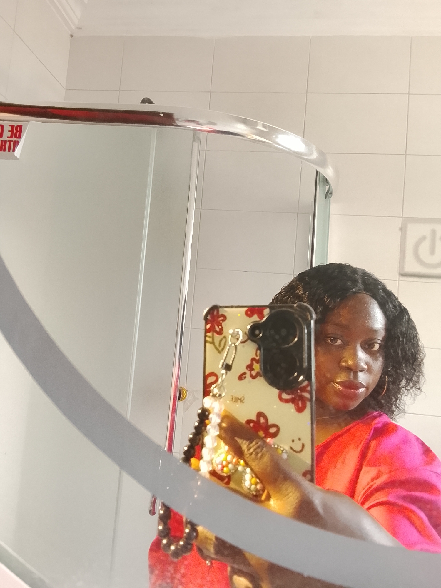A person is taking a mirror selfie in what appears to be a bathroom with white tiled walls. The phone case has red floral designs and a beaded charm. The person is wearing a bright pink shirt. The reflection in the mirror shows a shower stall with a curved, chrome-like frame. A power button symbol is visible on the tiled wall. This location is in Gwarinpa, Nigeria.