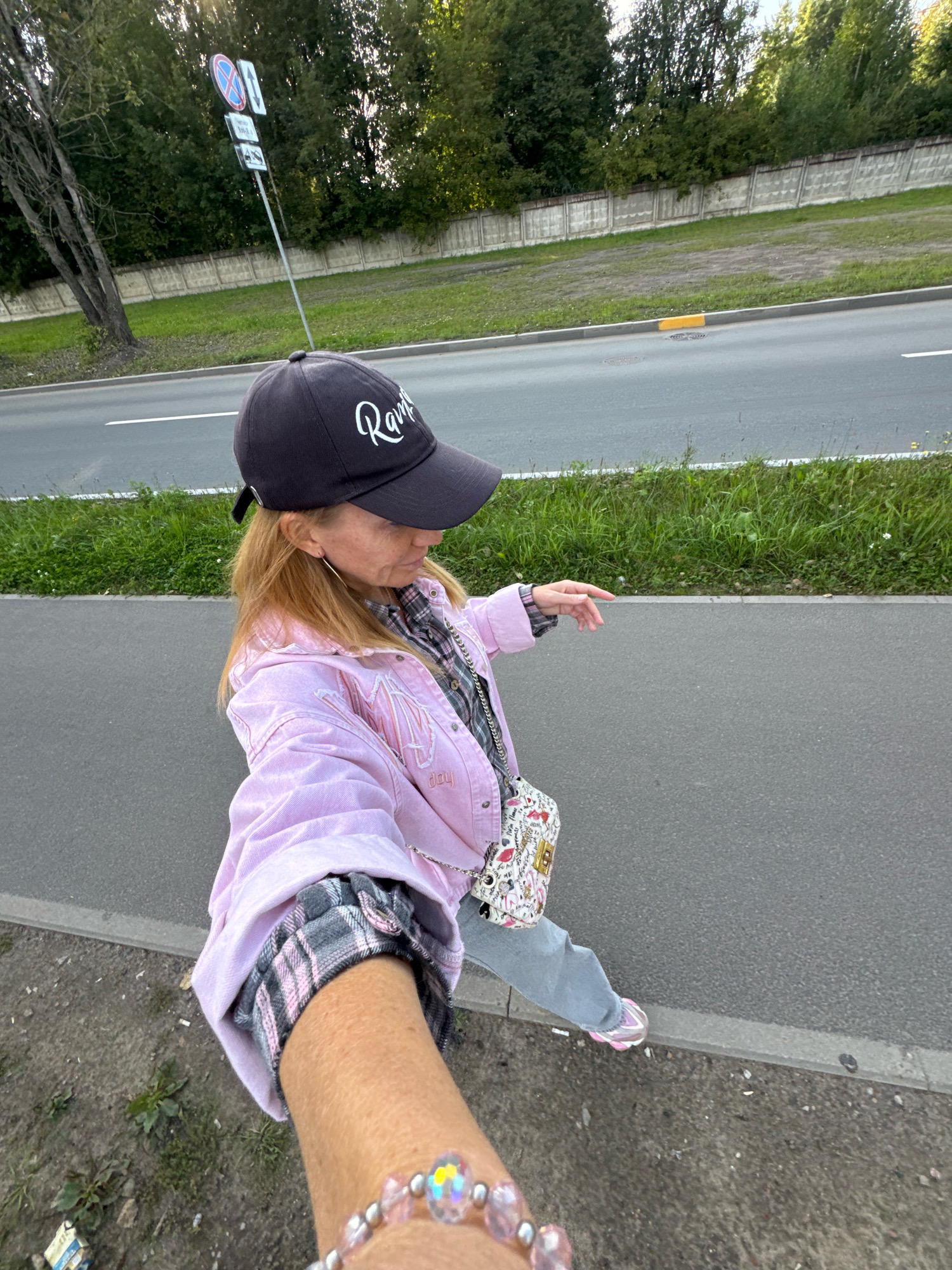 A woman, wearing a pink denim jacket, grey plaid shirt, blue jeans, a black baseball cap, and pink and white sneakers, is walking on a sidewalk next to a road. The woman has blonde hair and is wearing a beaded bracelet. She is also carrying a small, patterned crossbody bag. In the background, there is a concrete wall, trees, and a road with traffic signs. The scene appears to be in a suburban or semi-urban area.