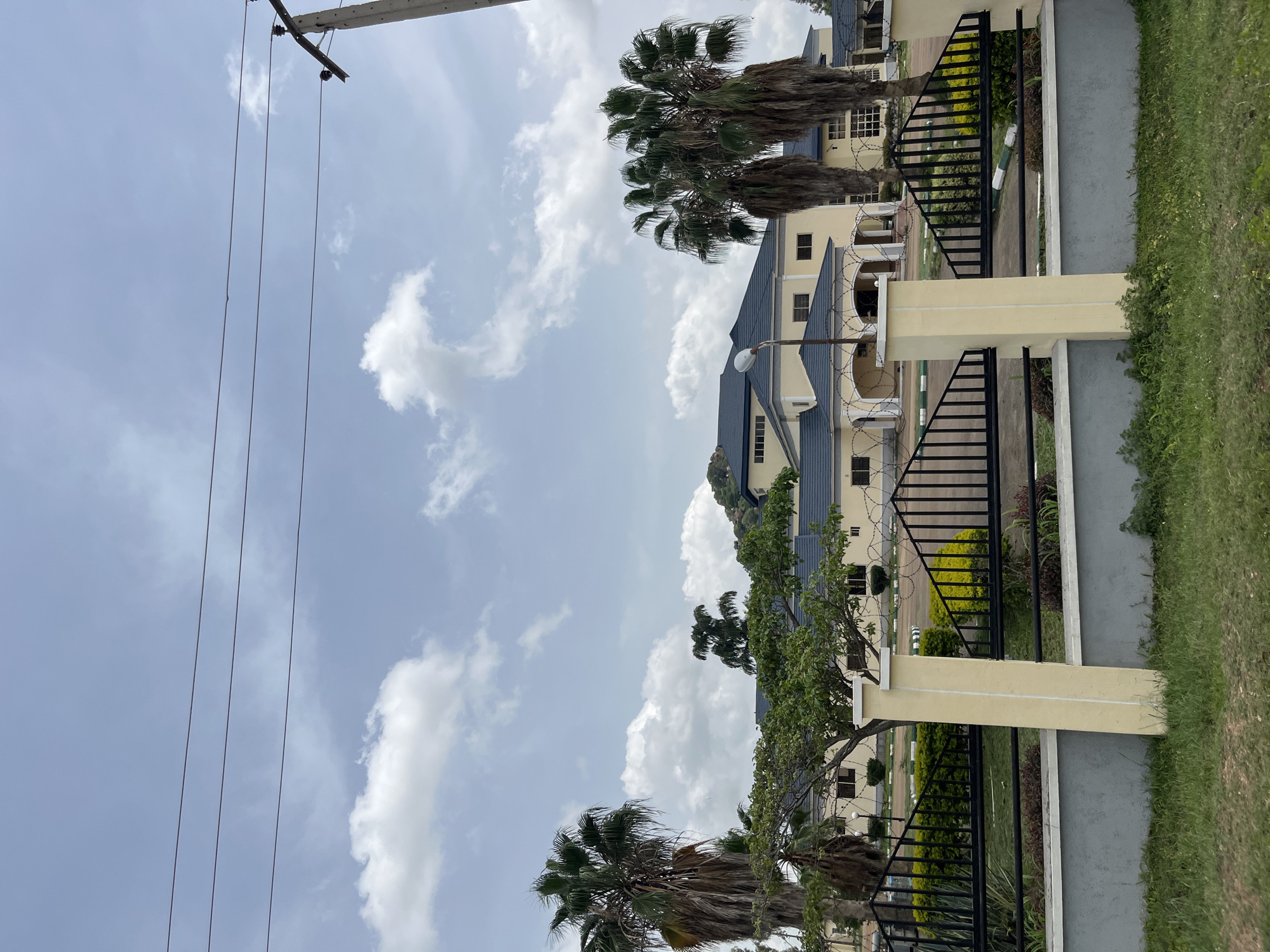The image depicts a large, light yellow building with a dark blue tiled roof, featuring multiple arched entrances and large windows. The building is surrounded by a black metal fence with decorative spikes and barbed wire, supported by light yellow concrete pillars. Tall palm trees are visible on either side of the building, and there are also green trees and shrubs in the foreground and around the property. The sky is blue with scattered white clouds. Electrical wires are strung across the top of the frame, supported by a concrete pole on the right. The scene appears to be a government building or a large private residence in a semi-urban or suburban area of Bukuru, Nigeria. There are no people visible in the image. The overall impression is one of security and grandeur.