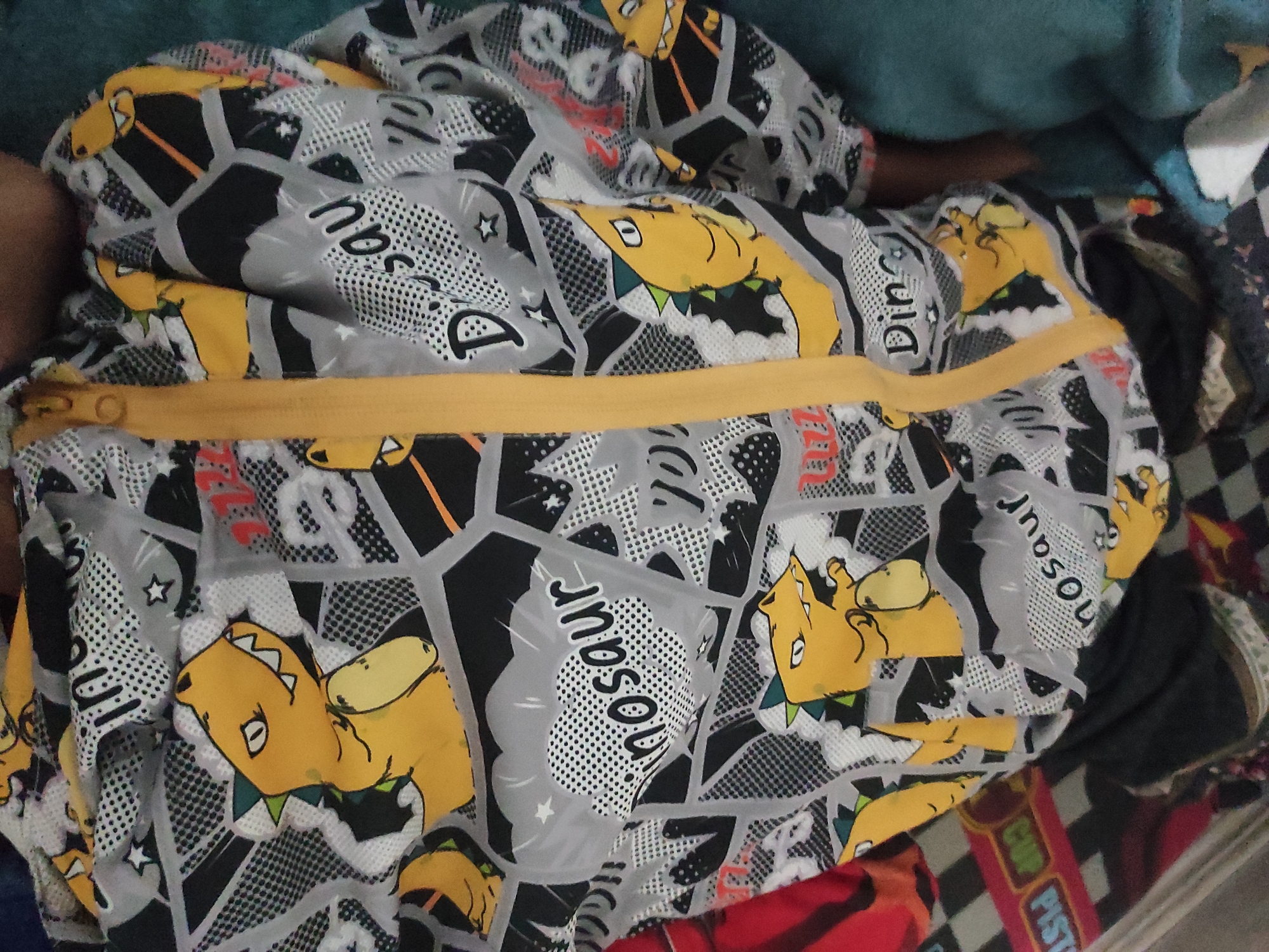 A close-up shot shows a yellow zipper on a grey hooded jacket with a cartoon dinosaur pattern. The jacket is draped over a person. The background is blurred but appears to be a bed with a checkered blanket. The location is Sumedang, Indonesia.