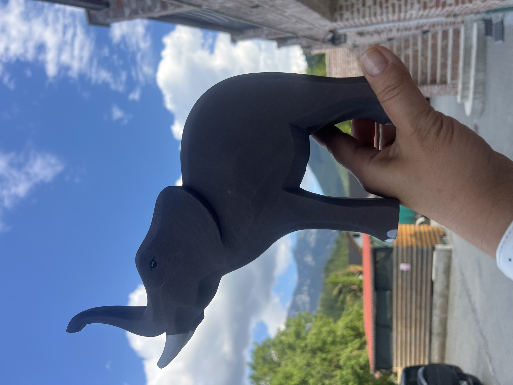 An outdoor medium shot captures a dark brown, stylized wooden elephant figurine held in the palm of a person's right hand. The elephant is depicted with its trunk raised upwards and one tusk is visible. The background features a bright blue sky with scattered white clouds. In the middle ground, lush green trees and a building with a red roof are visible, with a mountain range in the far distance. To the right, a brick building with stairs leading up is present. The foreground is dominated by the hand holding the elephant. The setting appears to be a village or rural area in an unspecified location.