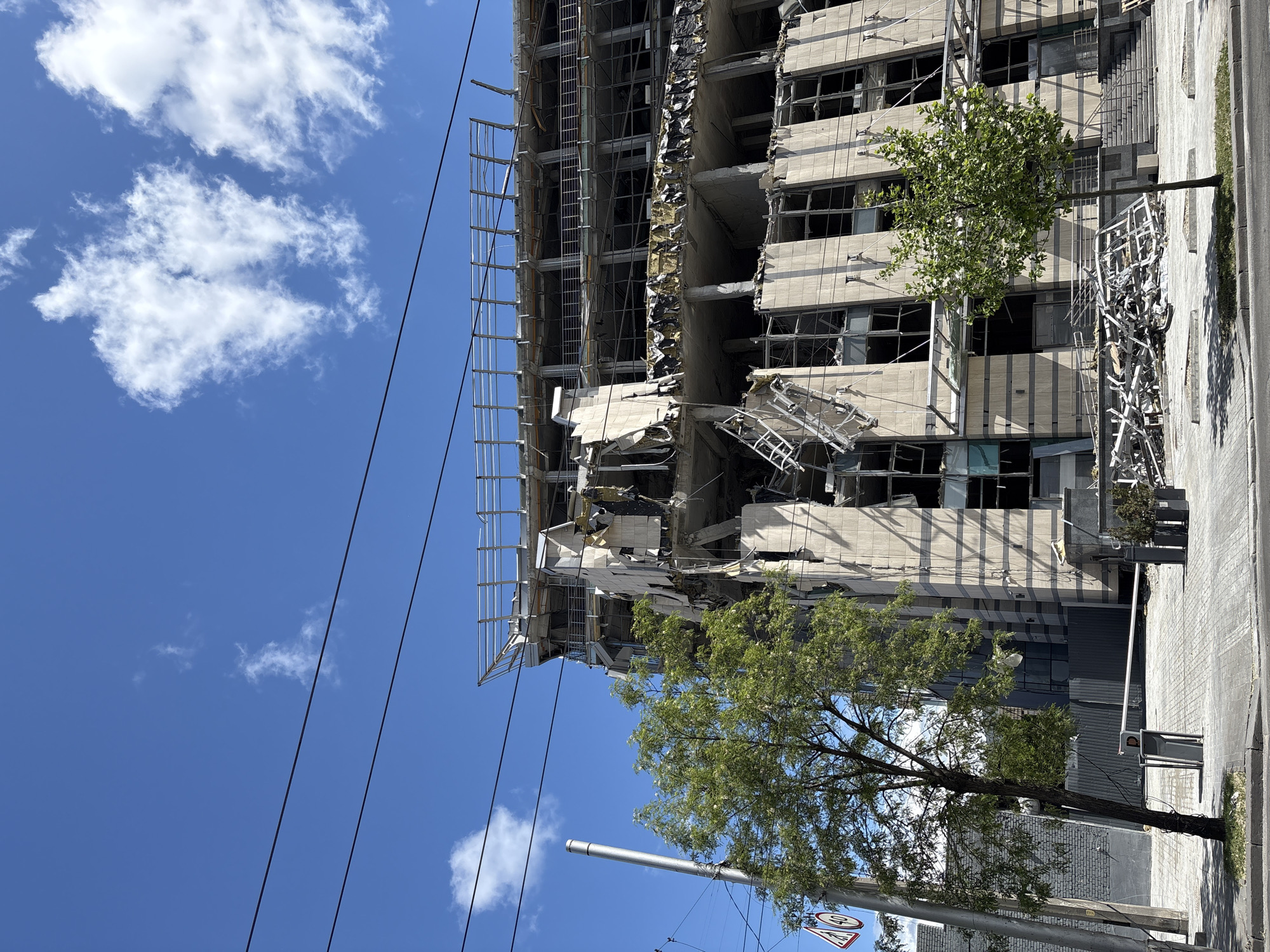 The image shows a damaged building in Kyiv, Ukraine. The building has multiple floors with broken windows and significant structural damage, including collapsed sections. Metal scaffolding is visible along the side of the building. The sky is blue with scattered clouds. Trees are visible near the building, and street-level infrastructure like utility poles and signage can be seen. The setting is urban, with a paved street alongside the building.