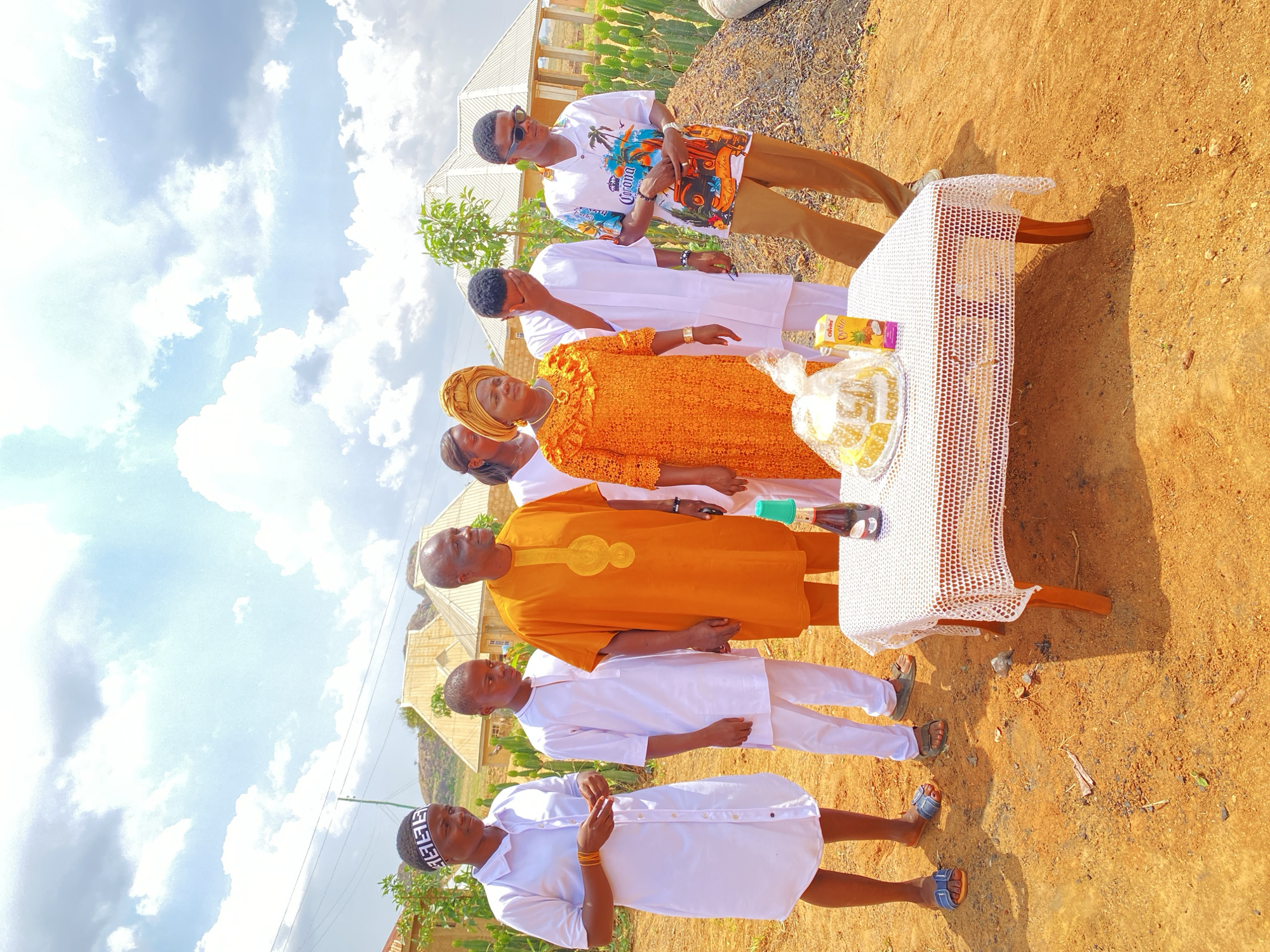 A group of seven people stands together outdoors in what appears to be Kuru Karama, Nigeria. One person is wearing a bright yellow dress and head wrap. Another individual is wearing a bright orange long-sleeved shirt and pants. Two people wear patterned shirts and khaki pants, while the others are dressed in white long-sleeved shirts and pants. They are gathered around a table covered with a white crocheted tablecloth. On the table, there are items including juice boxes and what seems to be wrapped food. The ground is a reddish-brown color with sparse vegetation. In the background, houses are visible under a sky filled with clouds.