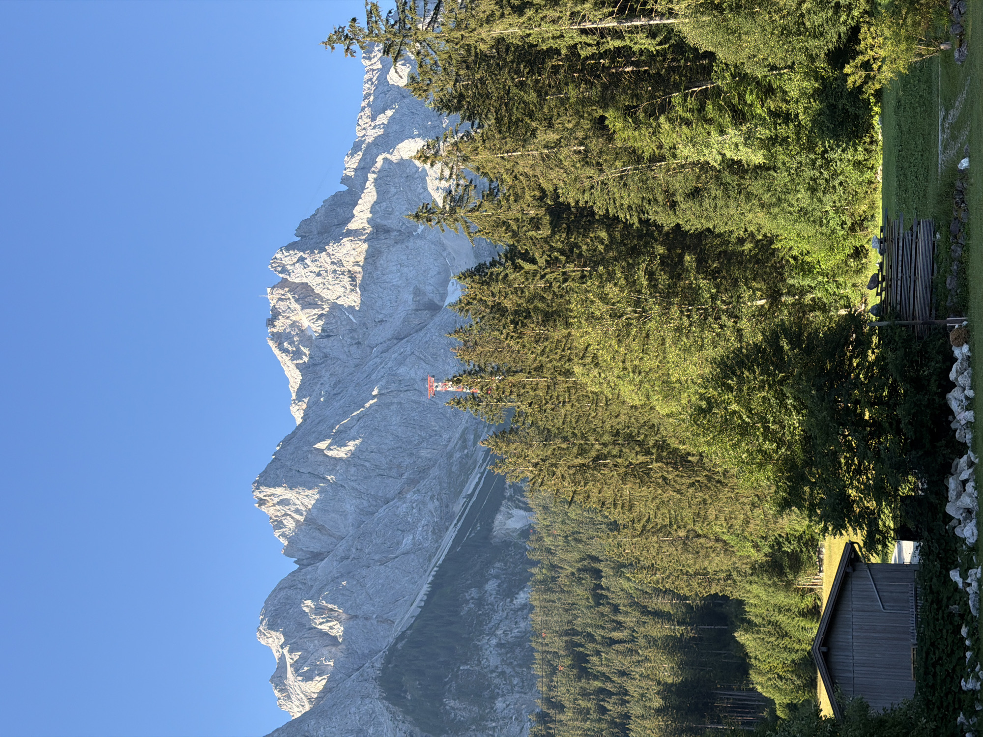 The image depicts a scenic mountain landscape with a clear blue sky. In the foreground, a wooden shed stands on the left, partially obscured by foliage and rocks. Adjacent to the shed, a grassy area with some wooden structures and benches is visible. Dense evergreen forests cover the mid-ground, leading up to a majestic mountain range. A red and white tower, possibly a ski lift or radio tower, stands out amongst the trees. The mountains are rugged and rocky, with some patches of snow or light-colored rock. The sunlight suggests it is either early morning or late afternoon, casting shadows and highlighting the textures of the terrain. The setting is likely Grainau, Germany, given the characteristic alpine scenery. There are no people visible in the image.