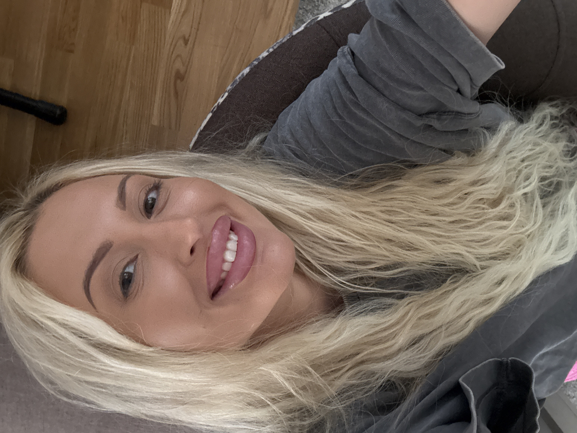 A woman with blonde hair and full lips smiles for a selfie while reclining on a sofa. Her face is close to the camera, and her blonde hair falls over her shoulders. She is wearing a gray sweatshirt. The background shows a wooden floor and a piece of furniture.