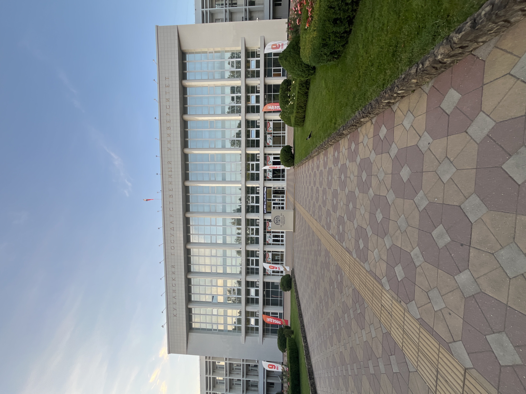 The image shows the main entrance to the Kuban State University in Krasnodar, Russia. The building is a large, modern structure with a facade of glass and light-colored stone. The words "КУБАНСКИЙ ГОСУДАРСТВЕННЫЙ УНИВЕРСИТЕТ" (Kuban State University) are inscribed in large letters above the entrance.
In front of the building is a wide, paved walkway made of geometric stone tiles in shades of brown, beige, and gray. A tactile paving strip, designed for visually impaired pedestrians, runs along the left side of the walkway. To the right of the walkway is a well-maintained lawn with manicured hedges and flowering bushes. A few red and pink roses are visible in the foreground.
Several flags are displayed in front of the building. Some of them bear the number "9" and the word "МАЙ" (May), suggesting a connection to Victory Day celebrations. Another flag is red with white text, possibly indicating a student or university event. There is also a large informational stand with the number "105" and text that appears to celebrate the university's anniversary.
The sky above is a clear blue with a few wispy clouds. A Russian flag is visible on a flagpole atop the building. A single crow is perched on the lawn in the middle ground. The overall impression is one of a well-maintained university campus on a clear day.