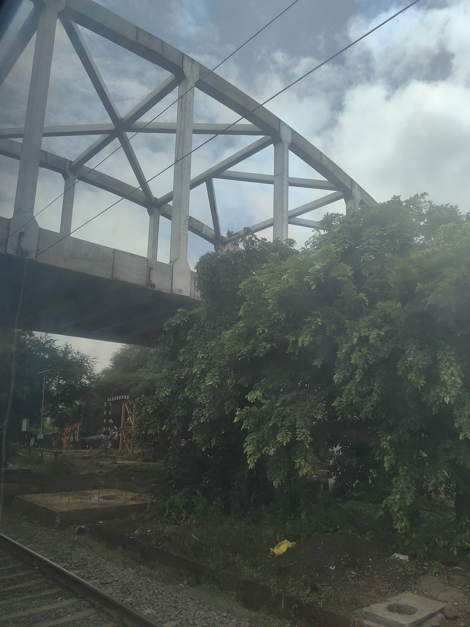 The image depicts a railway track running diagonally from the lower left corner towards the center. Overhead, a large concrete bridge with a complex truss structure spans across the frame. Dense green foliage of trees obscures the lower portion of the bridge and the surrounding area, suggesting a lush environment. In the background, under the bridge, several construction elements are visible, including what appears to be an orange construction vehicle and a wooden structure with black and white markings, possibly a signal or marker. The ground is uneven and appears to be a mix of dirt, gravel, and some construction debris. A yellow plastic bag is visible on the ground amongst the vegetation. The sky above is cloudy with patches of blue visible through the clouds. The overall scene suggests an outdoor location, possibly on the outskirts of Kamtara, India, where infrastructure development or maintenance might be underway near a railway line.