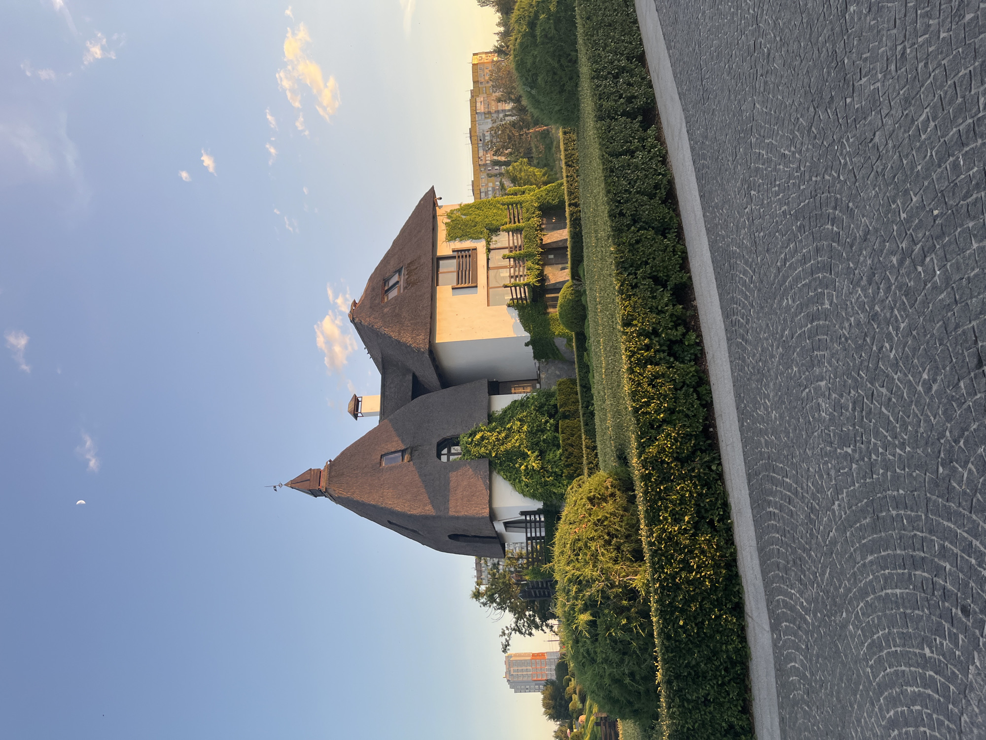 The image shows a distinctive house with a thatched roof, featuring multiple gables and a tall, spire-like turret. The house is surrounded by meticulously maintained greenery, including hedges and sculpted bushes. A cobblestone pathway curves in the foreground, leading towards the house. In the background, several multi-story residential buildings are visible, indicating an urban or suburban setting. The sky is clear with a few wispy clouds and a crescent moon is present. The lighting suggests it is either early morning or late afternoon. This house is located at Dnipro, Ukraine, within the neighborhood known as "Dnipro-Donbas" or "Dnipro-Donbas Housing Complex".