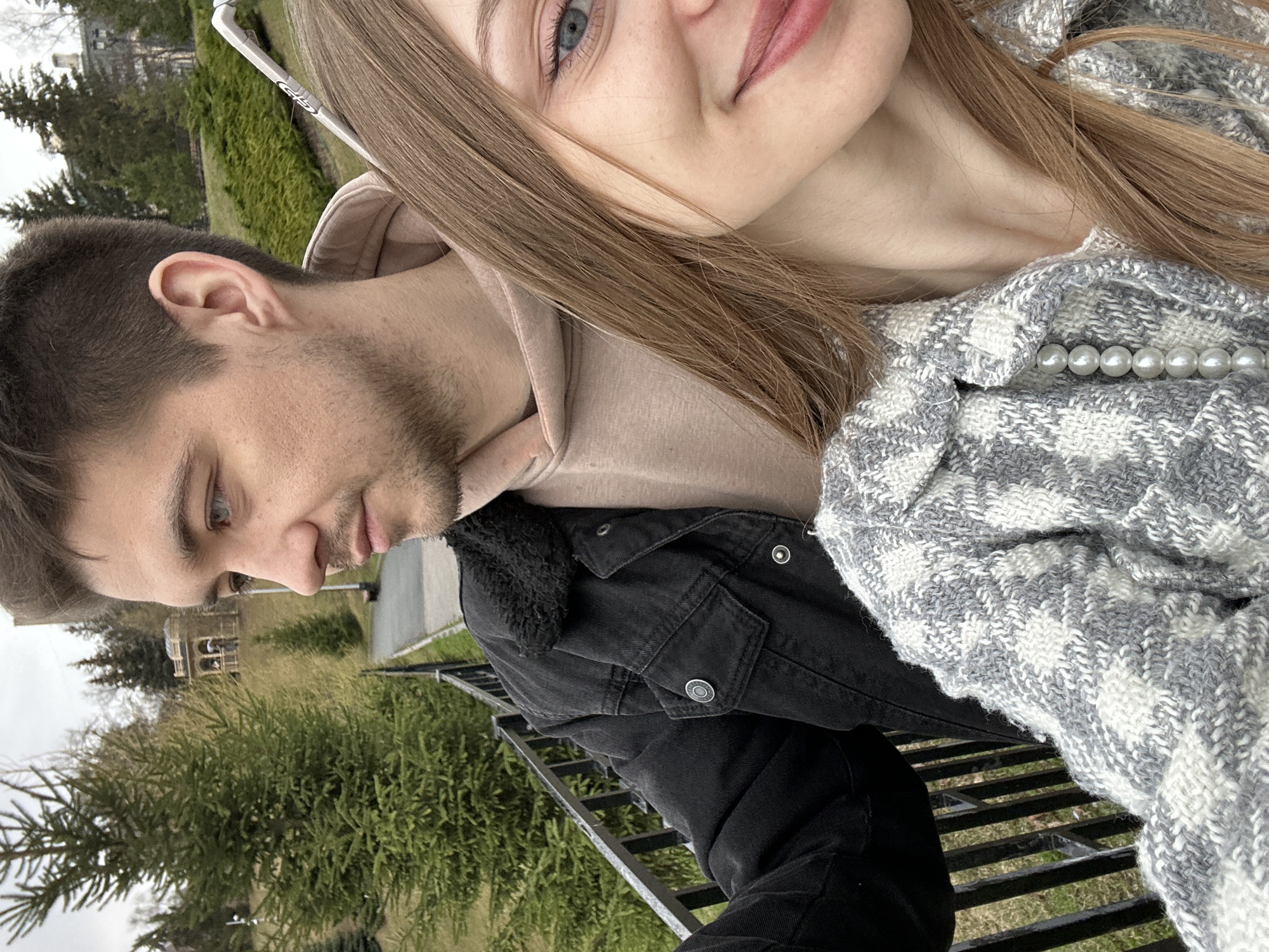 This outdoor image features a young man and woman in what appears to be a park or garden setting in Kazan, Russia. The shot is composed as a close-up, angled sideways.
On the right, a young woman is partially visible from her nose upwards. She has long, light brown hair, striking blue eyes, and pink-tinted lips, captured with a soft, gentle smile. She is wearing a grey and white patterned jacket, possibly tweed or knit, which is elegantly detailed with a visible string of white pearls near the collar. A thin white bar or frame, possibly part of sunglasses or a railing, is seen behind her hair.
On the left, a young man is visible from his head to his torso, looking downwards with a neutral expression. He has short light brown hair and light stubble on his face. He is dressed in a light brown hooded sweatshirt layered under a black denim jacket featuring a dark, possibly sherpa, collar and metallic buttons.
The background consists of green trees and bushes, suggesting lush outdoor scenery. A faint, ornate structure resembling a gazebo is visible in the distant background beyond the man. A black metal railing or fence is present in the foreground, separating the subjects from the environment. The lighting suggests an overcast sky during daytime, implying cool weather, consistent with their layered clothing. There is no visible text in the image. The overall mood is relaxed and candid.